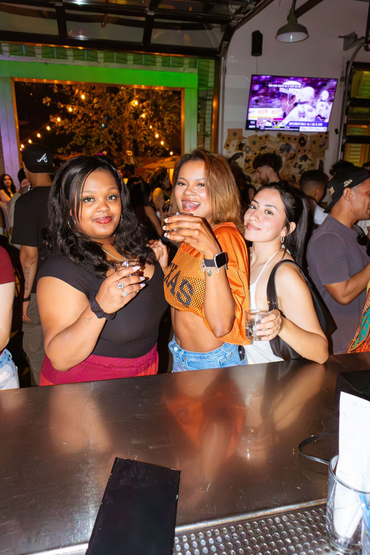 Group of friends enjoying drinks at a crowded bar with TVs and festive lighting.