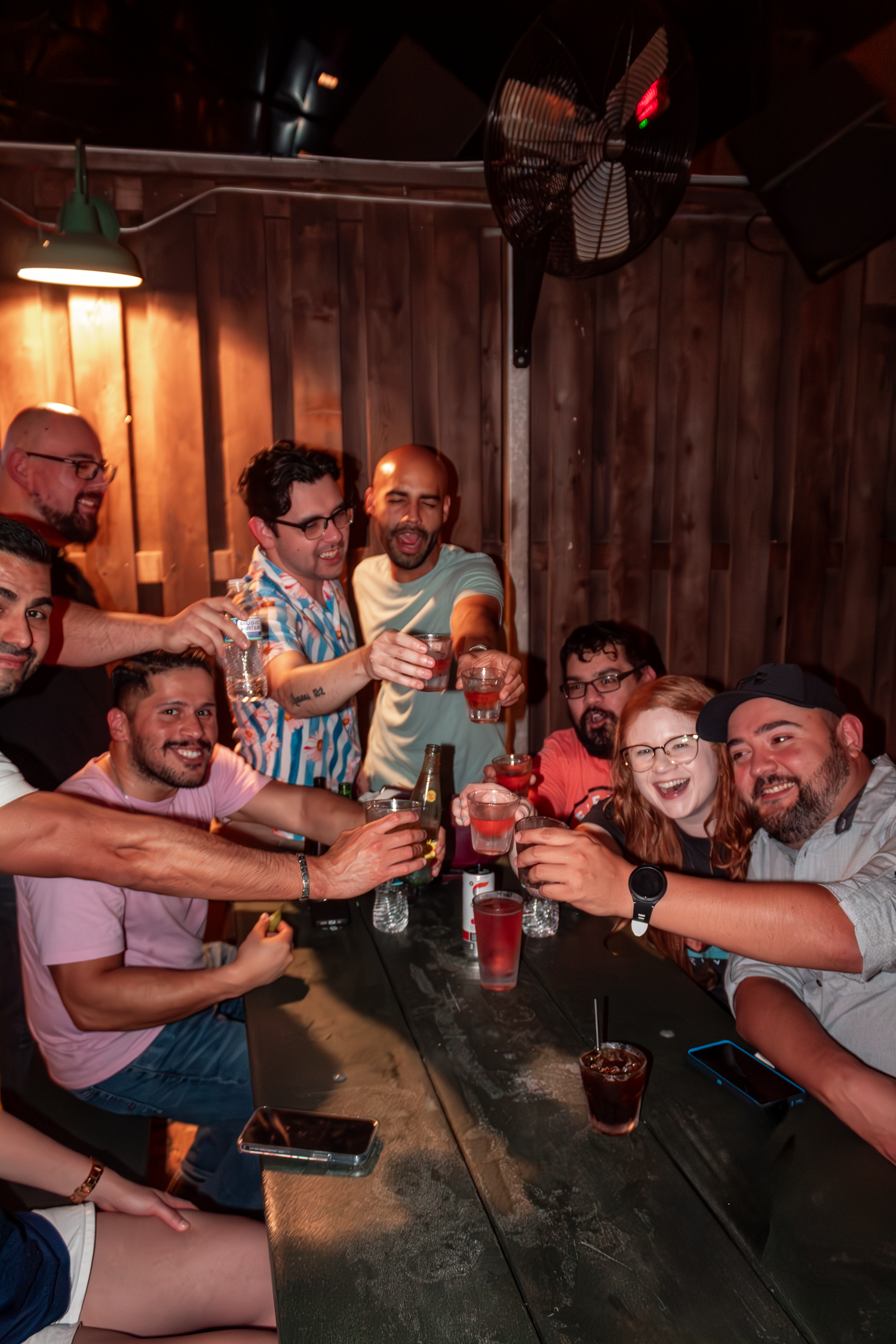 Group of friends toasting drinks around a wooden table in dimly lit bar setting at Camp 1604 in San Antonio, Texas.