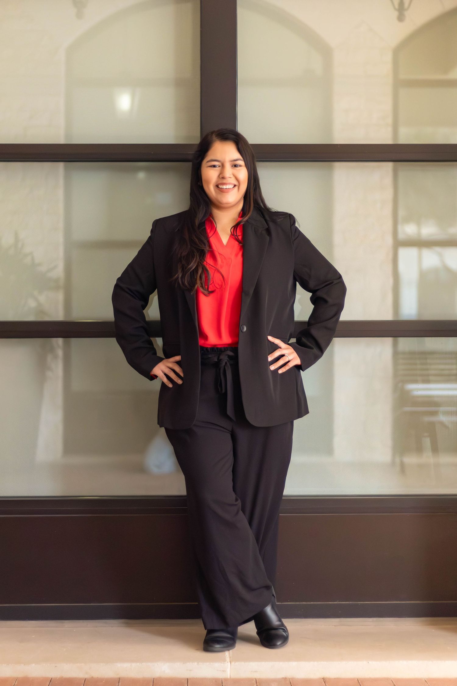 Professional woman in black suit and red blouse poses confidently against architectural background.