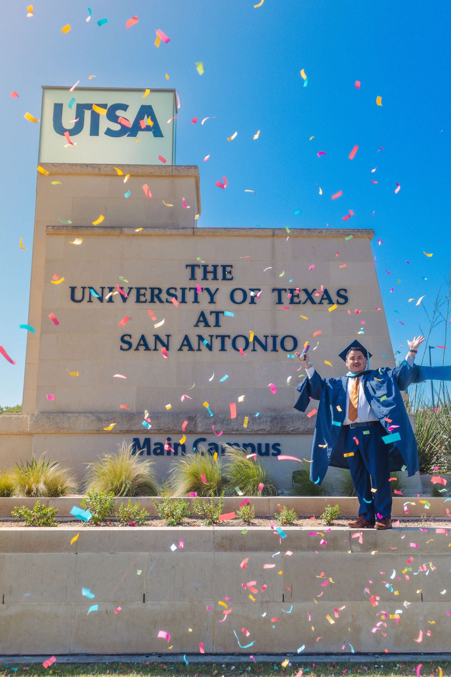 Colorful confetti falls around the main entrance sign of UTSA (The University of Texas at San Antonio) on a sunny day.