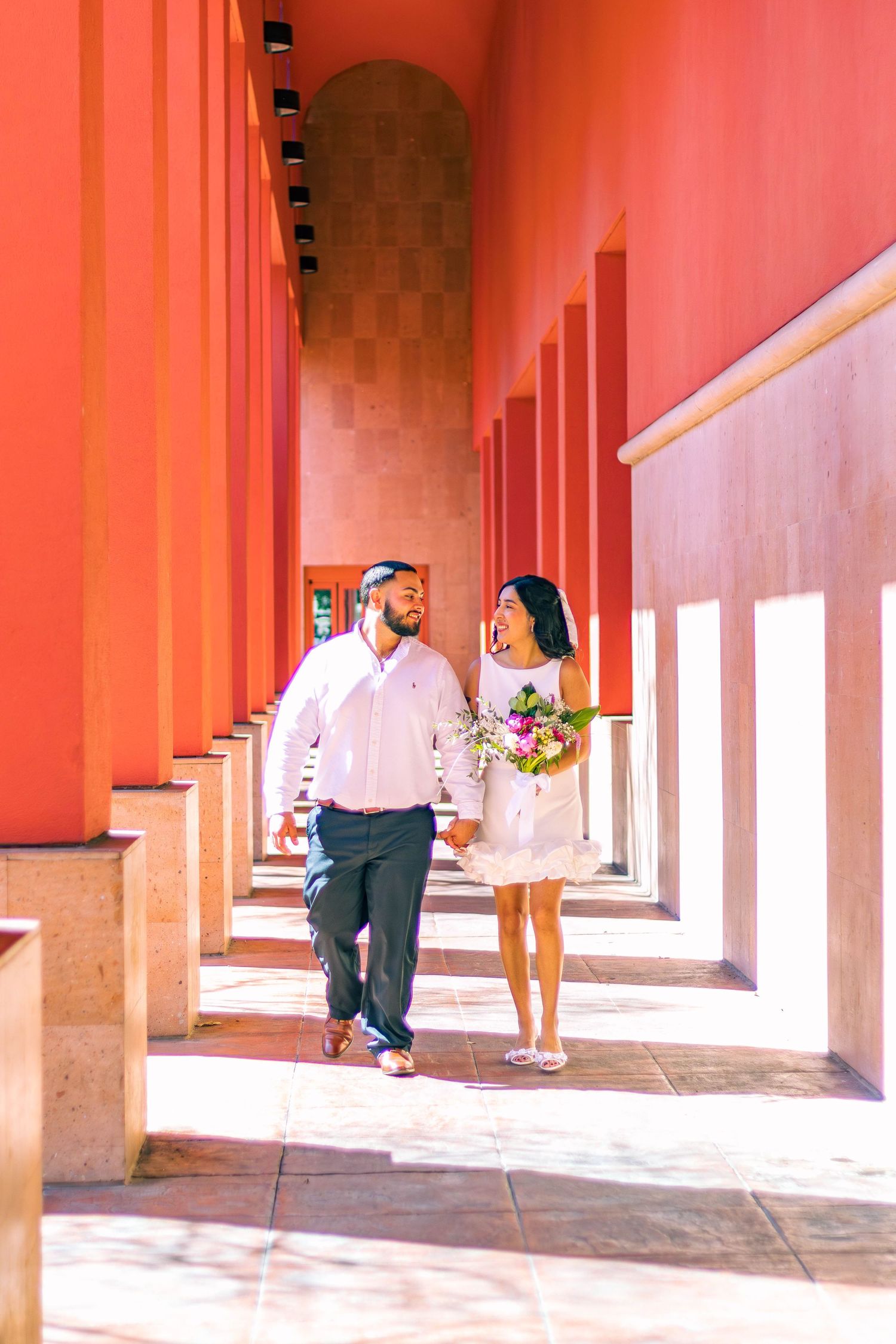 A couple strolls through a coral-colored colonnade with white pillars while holding a floral bouquet.