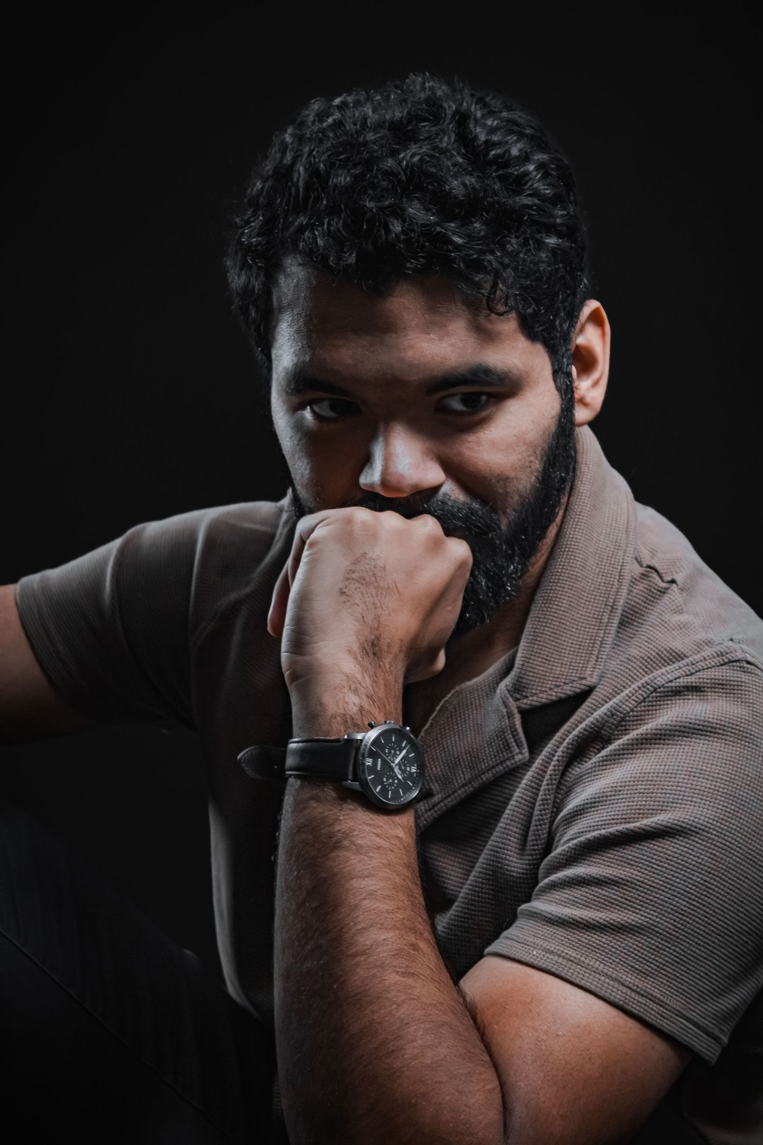 Man in brown shirt poses thoughtfully against dark background wearing watch.