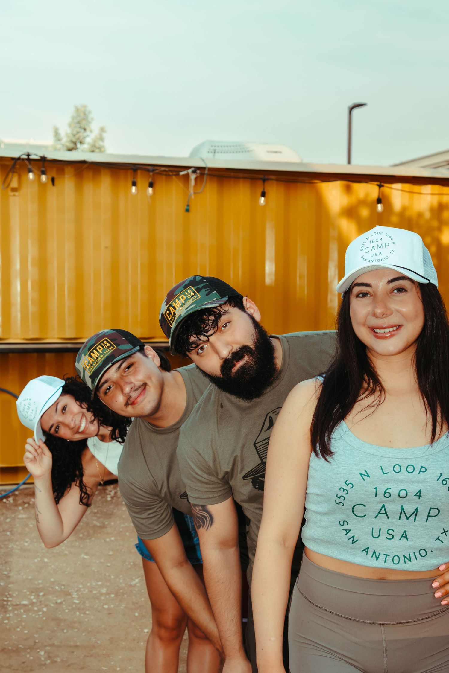 Group of friends in baseball caps lined up playfully against an orange shipping container background at Camp 1604 in San Antonio, Texas