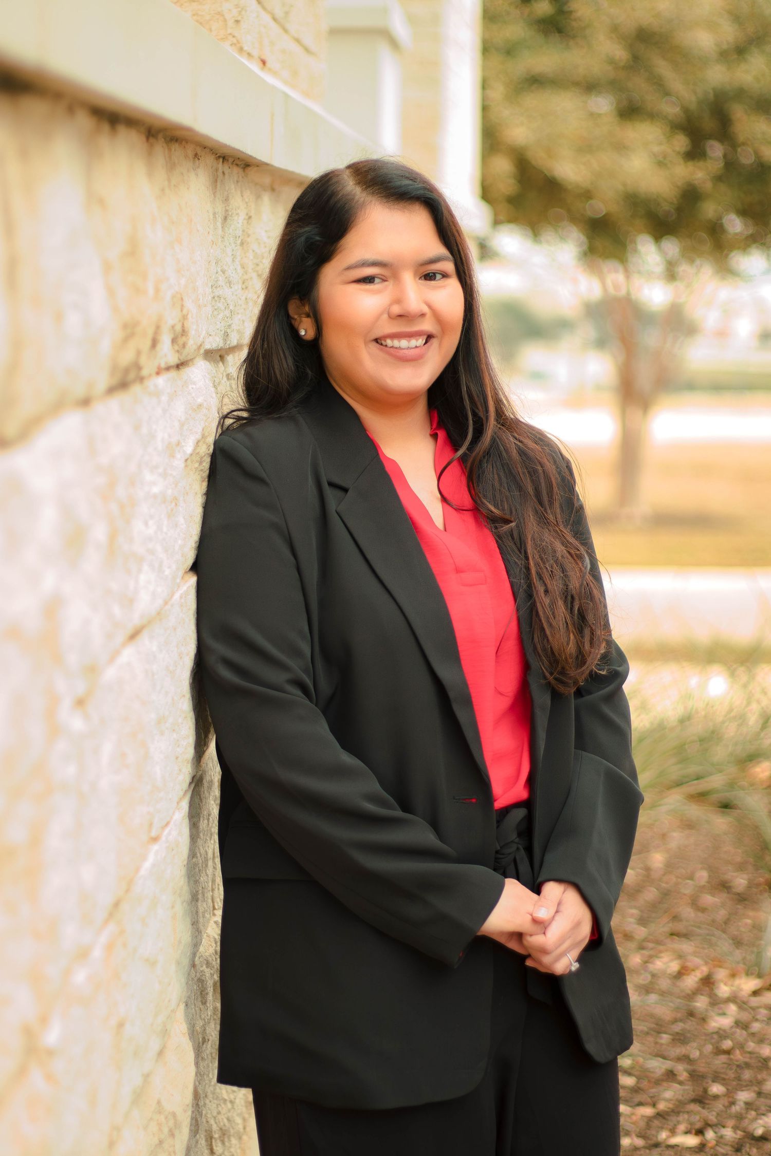 Professional headshot of woman in black blazer and red blouse against outdoor wall.