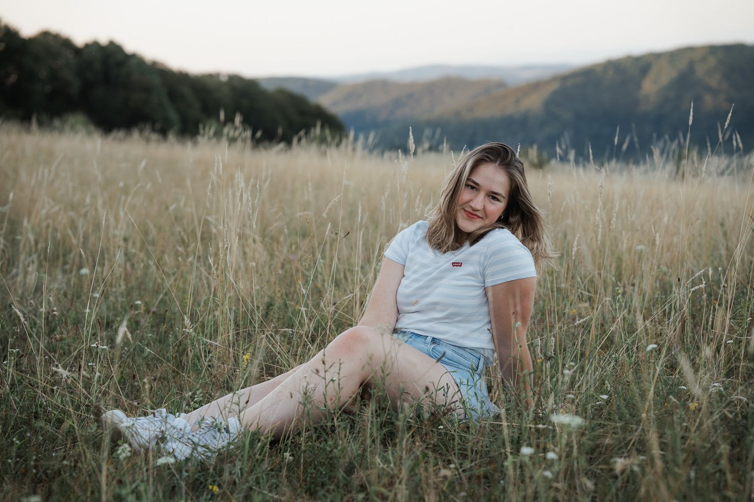 Mädchen in einem weissen T-Shirt und Jeans-Shorts sitzt auf einer goldenen Wiese bei Sonnenuntergang mit sanften Hügeln im Hintergrund.