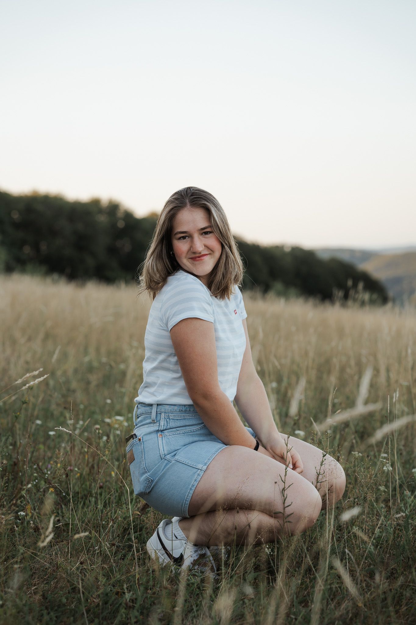 Mädchen in einem weissen T-Shirt und Jeans-Shorts sitzt auf einer goldenen Wiese bei Sonnenuntergang mit sanften Hügeln im Hintergrund.