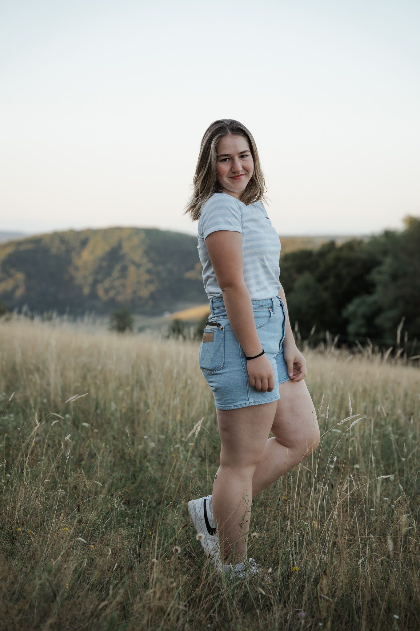 Mädchen in einem gestreiften T-Shirt und Jeans-Shorts steht auf einer goldenen Wiese bei Sonnenuntergang mit sanften Hügeln im Hintergrund.
