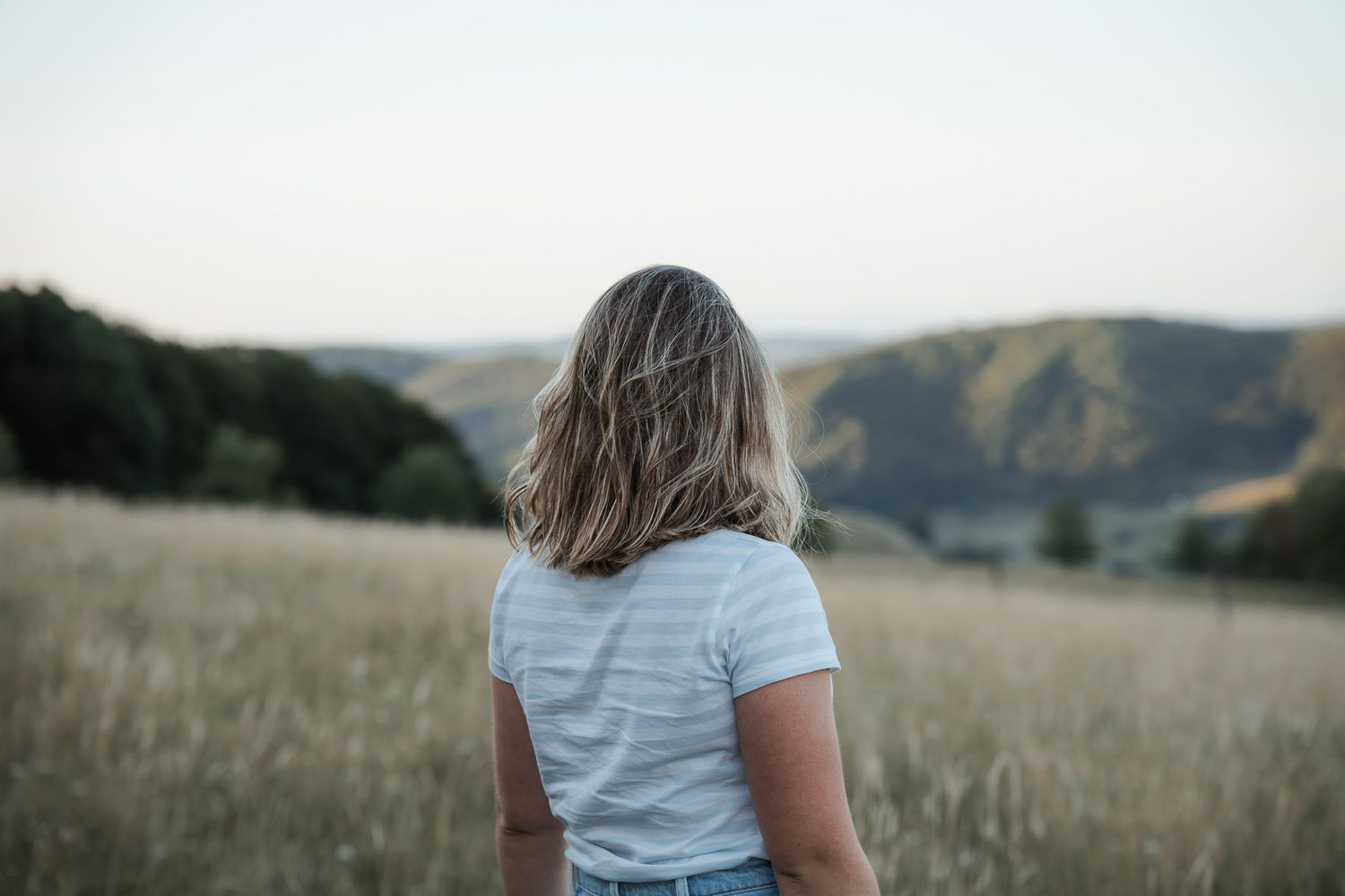 Mädchen in einem gestreiften T-Shirt und Jeans-Shorts steht auf einer goldenen Wiese bei Sonnenuntergang mit sanften Hügeln im Hintergrund.
