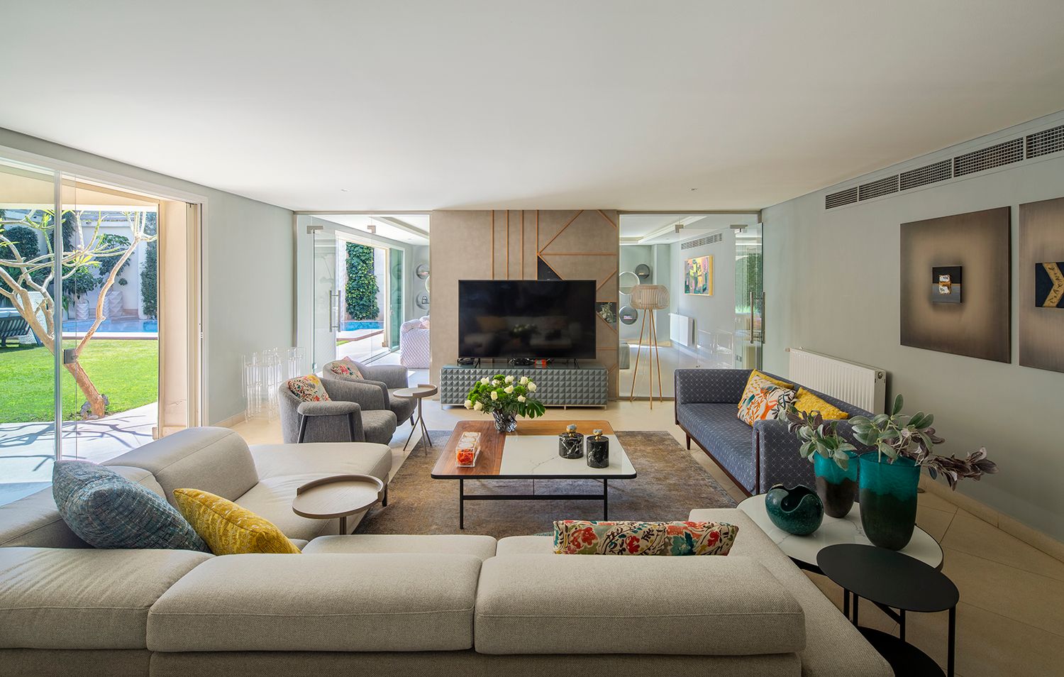 Modern living room with gray sectional sofa, mounted TV, and large windows overlooking outdoor space.
