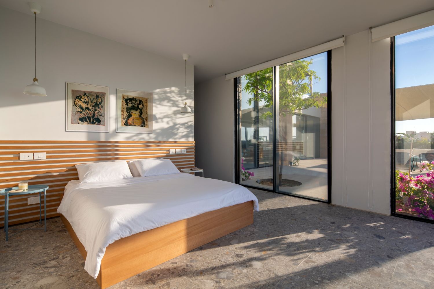 Minimalist bedroom with wooden bed frame, floor-to-ceiling windows, and natural light casting shadows.