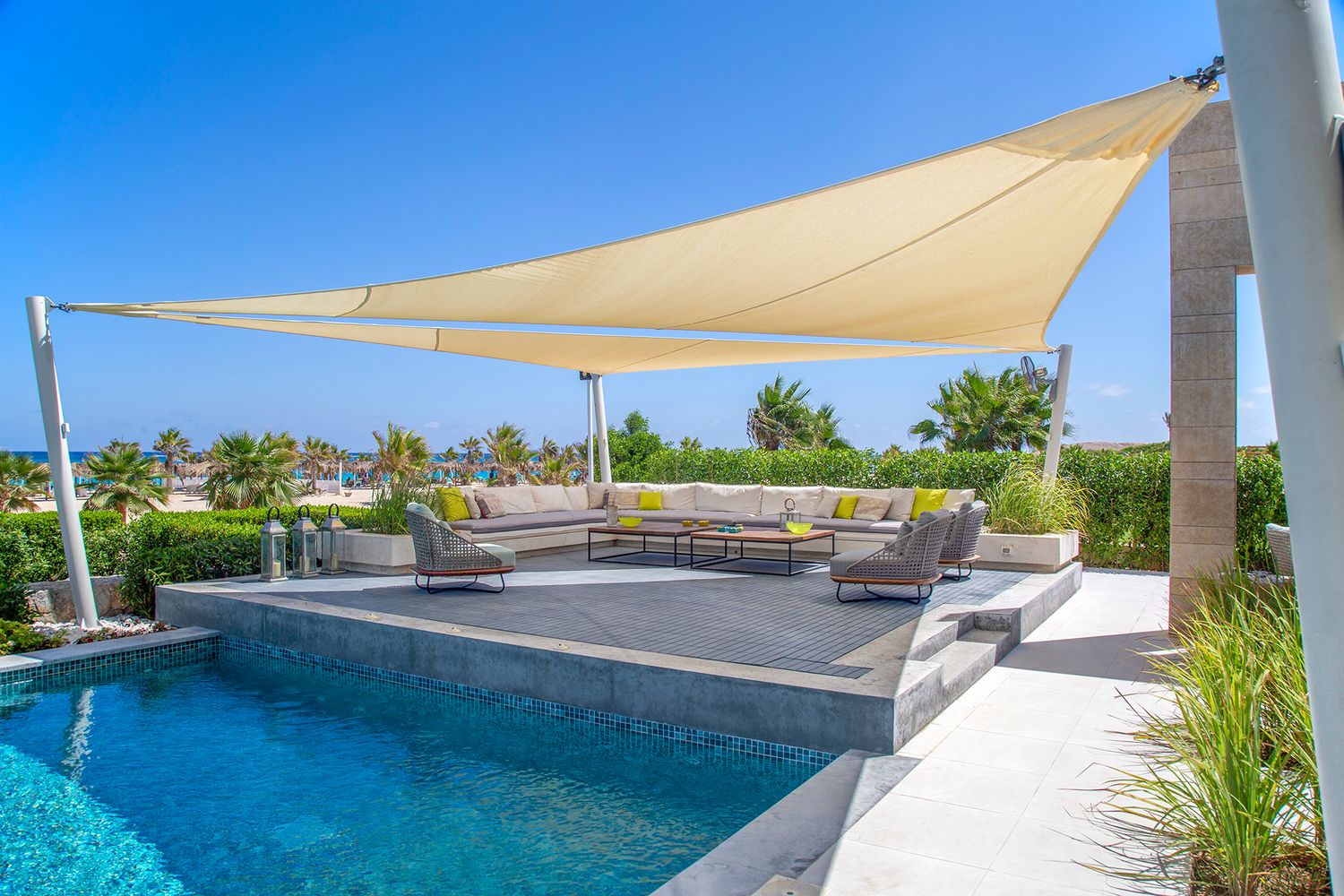 Modern poolside lounge area with beige sail shade and ocean view.
