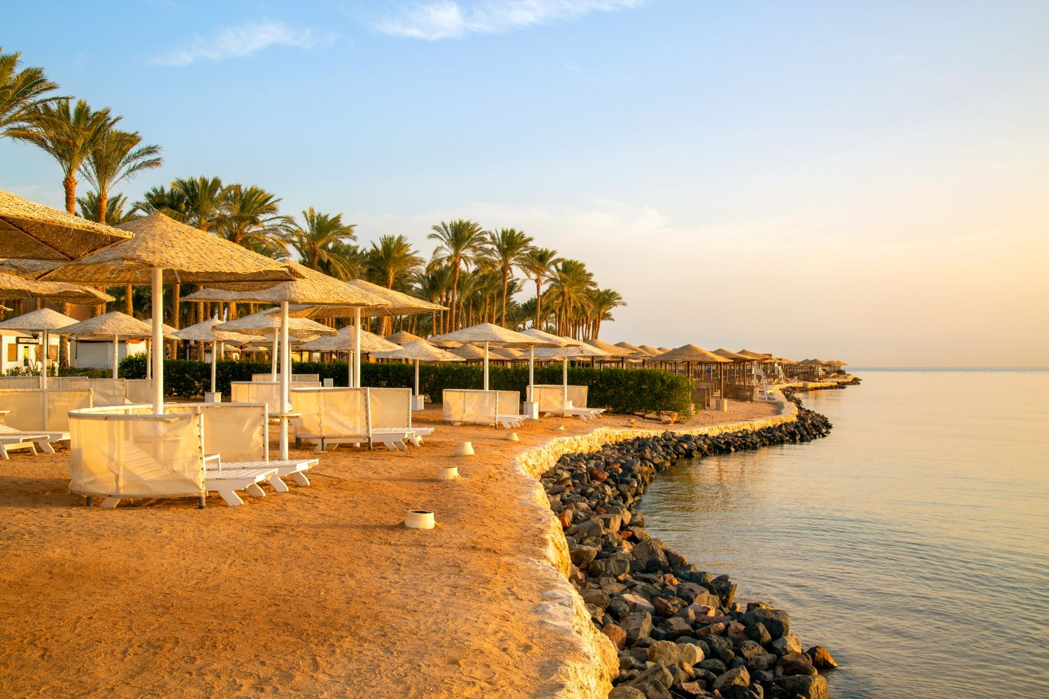Beachfront resort with palm trees and thatched umbrellas along a rocky coastline at sunset.