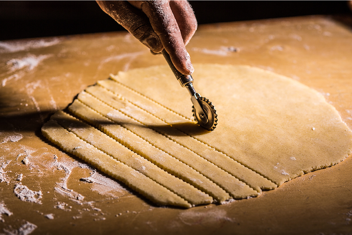 Close-up of cutting strips into rolled pasta dough on a floured surface.