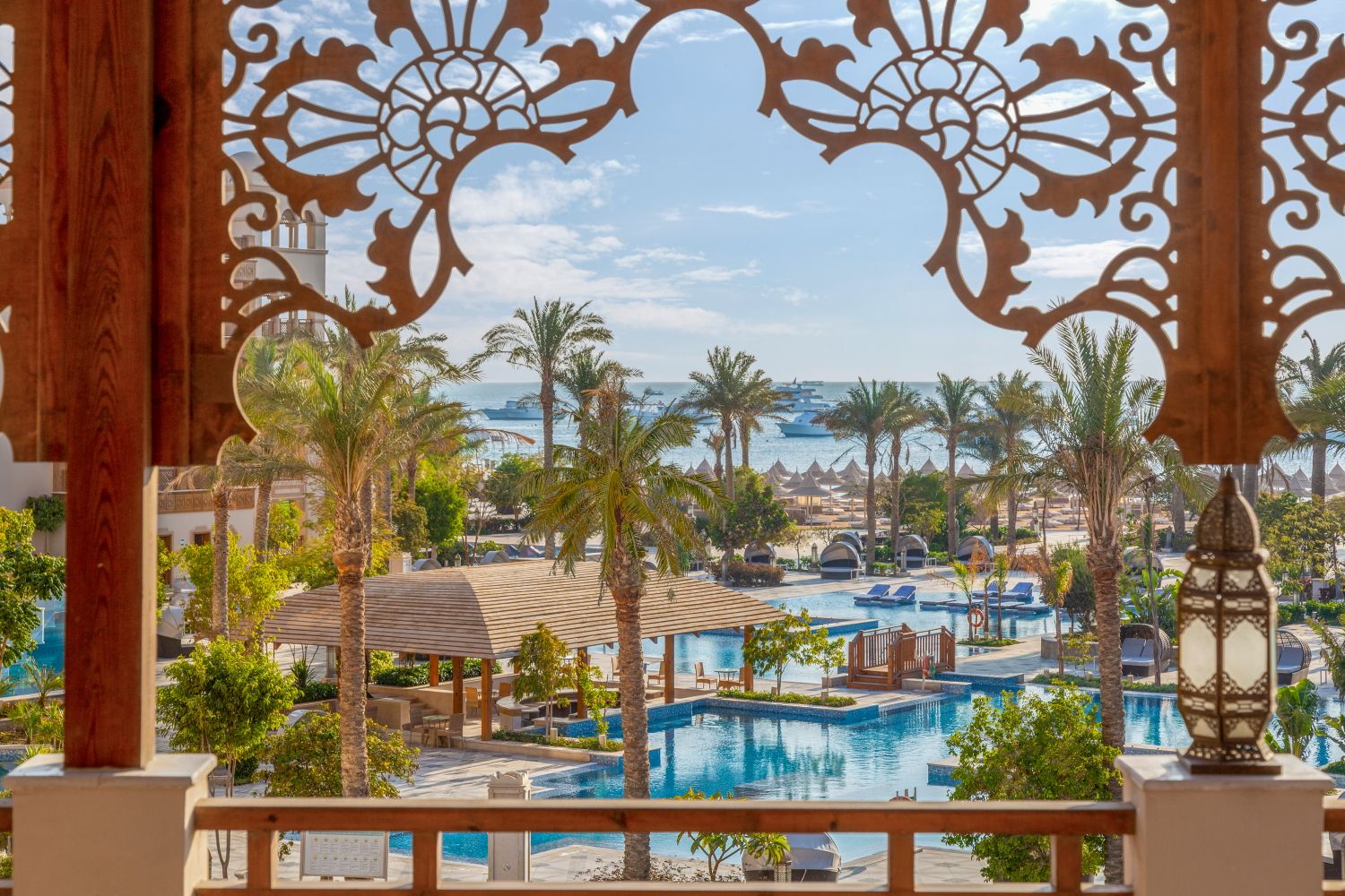 Resort pool view through ornate wooden archway with palm trees and ocean in background.