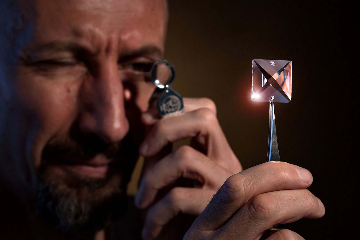 Person examining a small crystal part with a magnifying glass against a dark background with dramatic lighting.