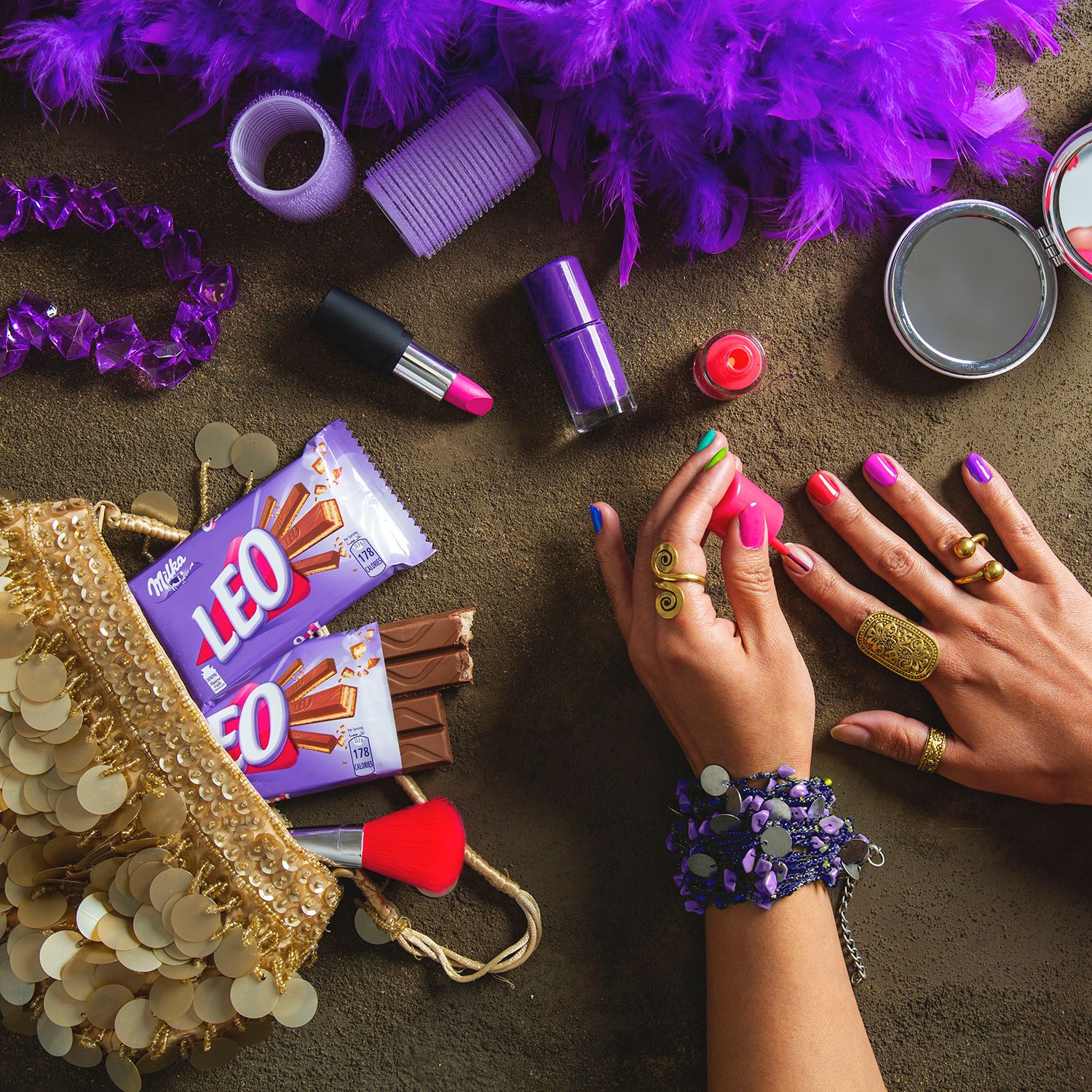 Flatlay of purple party decorations, feathers, Milka Leo chocolate bars and jewelry on dark background.