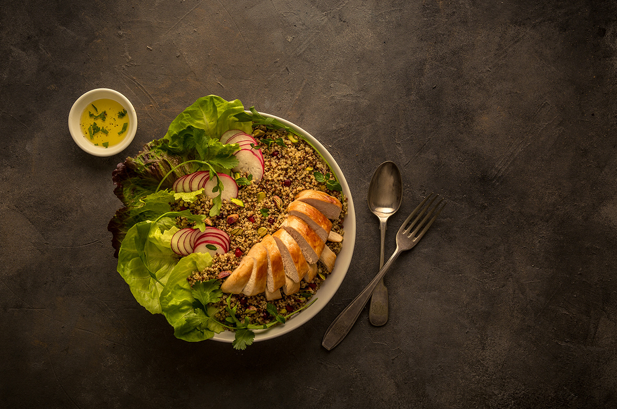 A healthy grain bowl with sliced chicken breast, fresh lettuce and quinoa served with dressing on dark background.