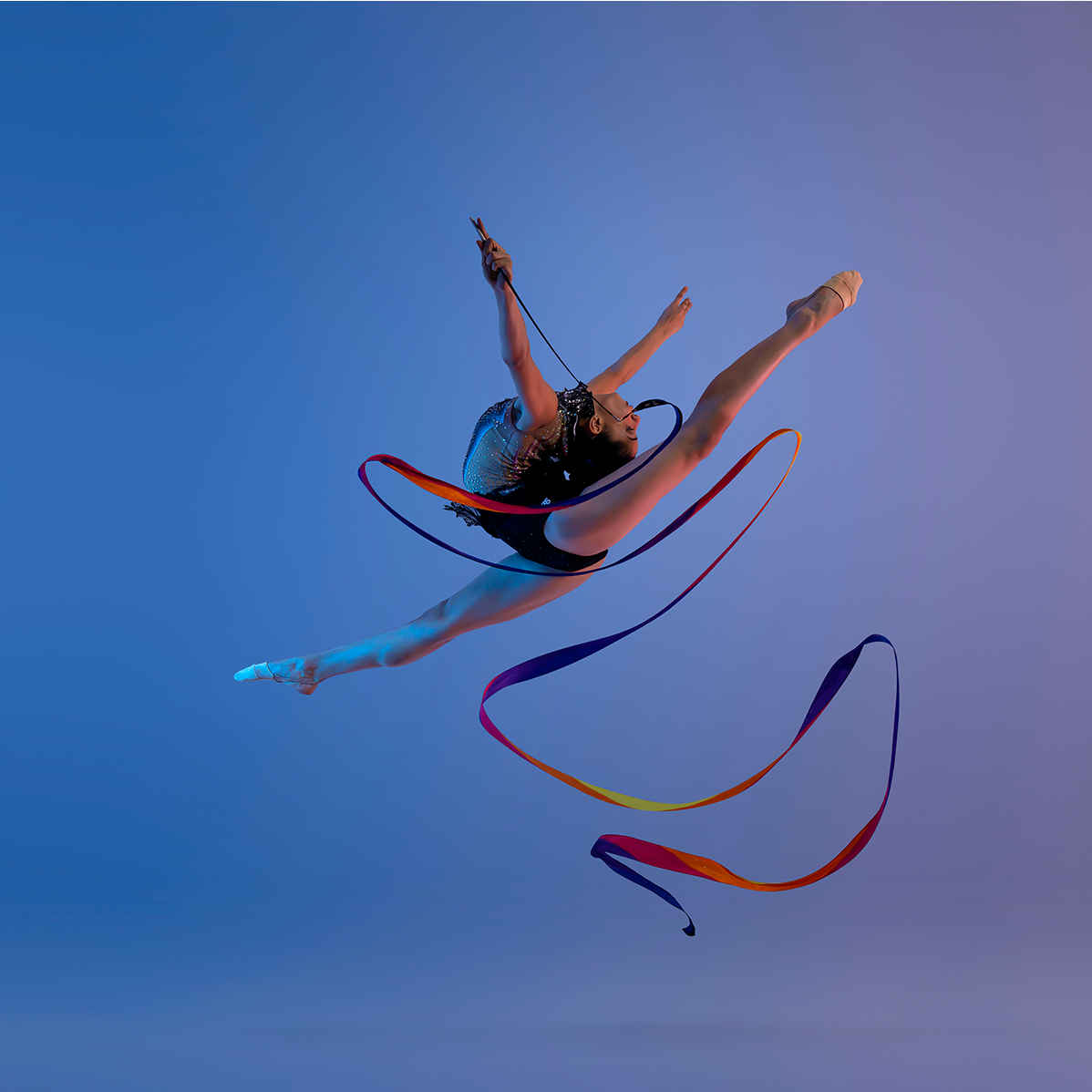Dynamic rhythmic gymnastics performance with ribbon creating flowing patterns against blue backdrop.