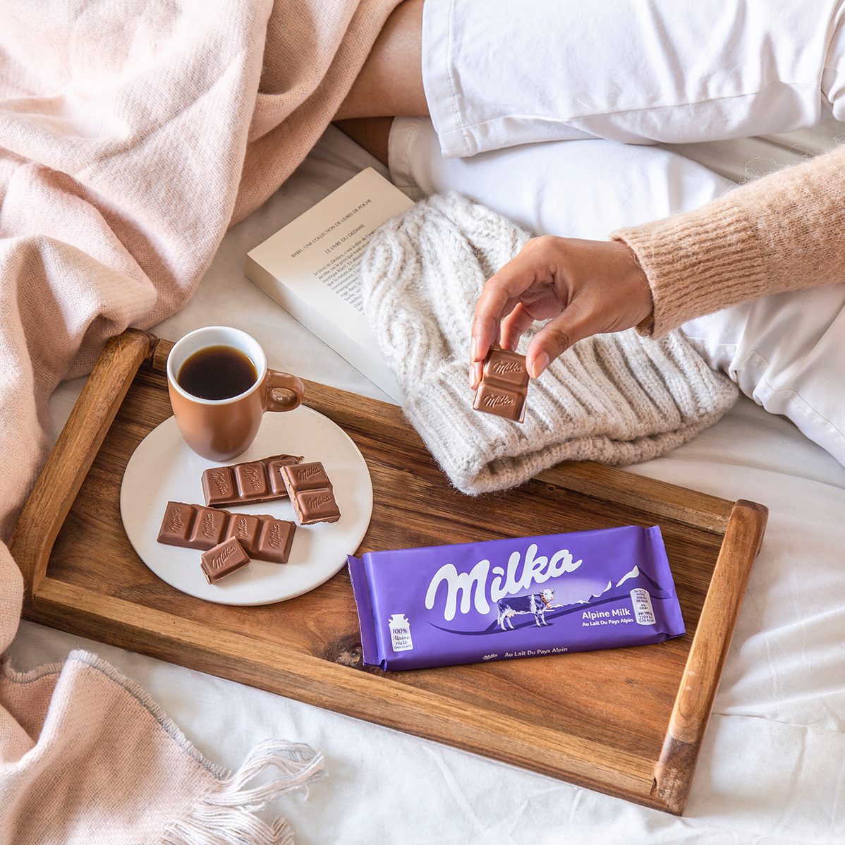 Cozy breakfast in bed setup with coffee, chocolates, and Milka bar on wooden tray.