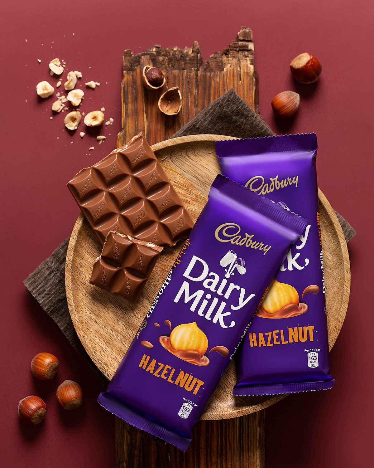 Cadbury Dairy Milk hazelnut chocolate bars with scattered nuts on wooden board against burgundy background.