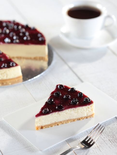 Slice of blueberry cheesecake with coffee on white plate against bright background.