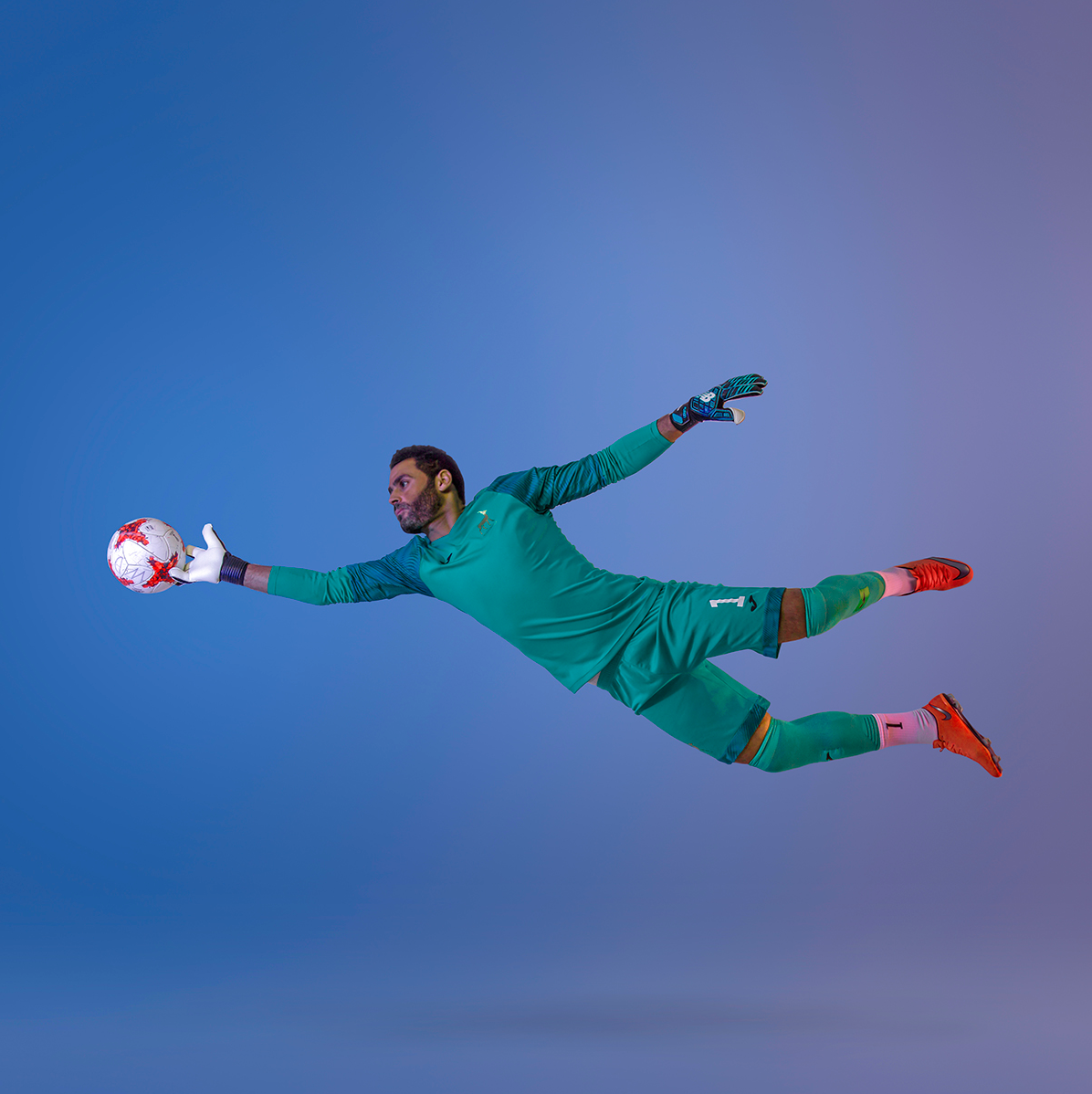 Soccer goalkeeper in green uniform making diving save against blue background.