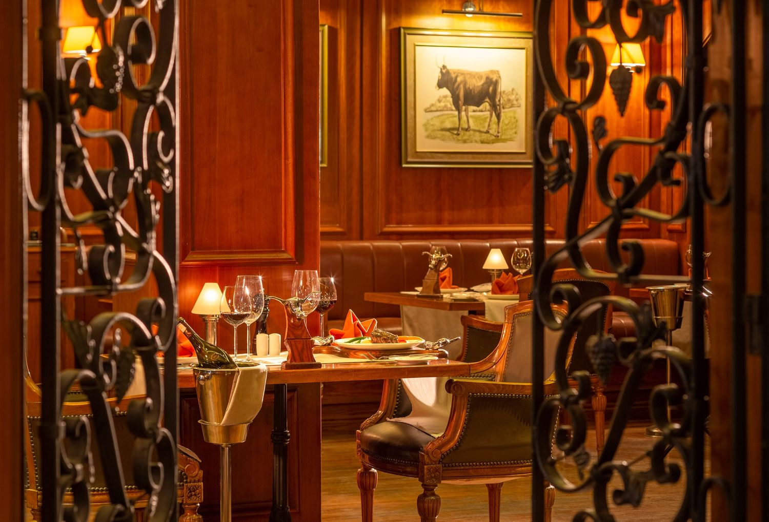 Elegant restaurant dining room viewed through ornate wrought iron gates with warm lighting and classic decor.