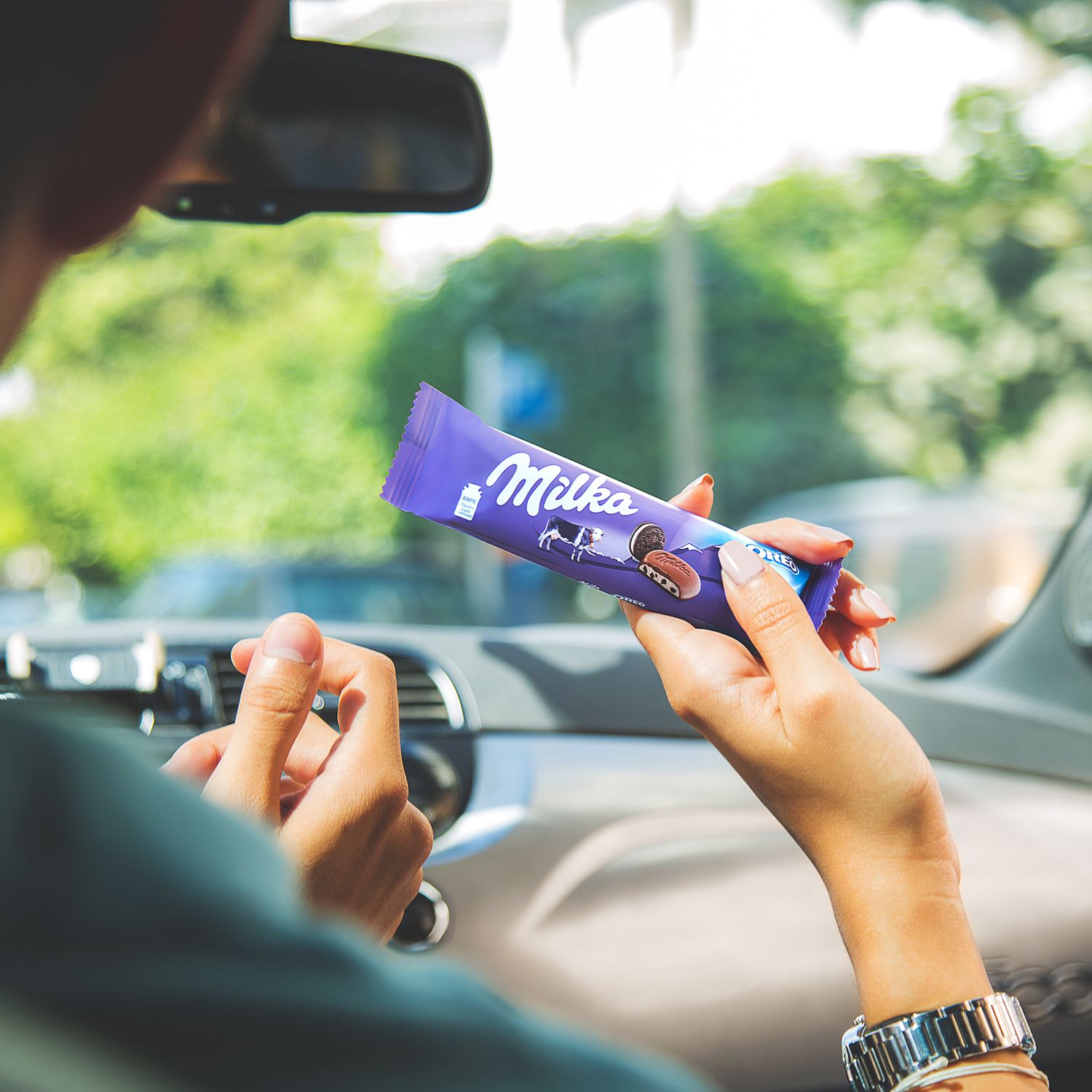 Hand holding Milka chocolate bar while driving, with green scenery visible through car window.