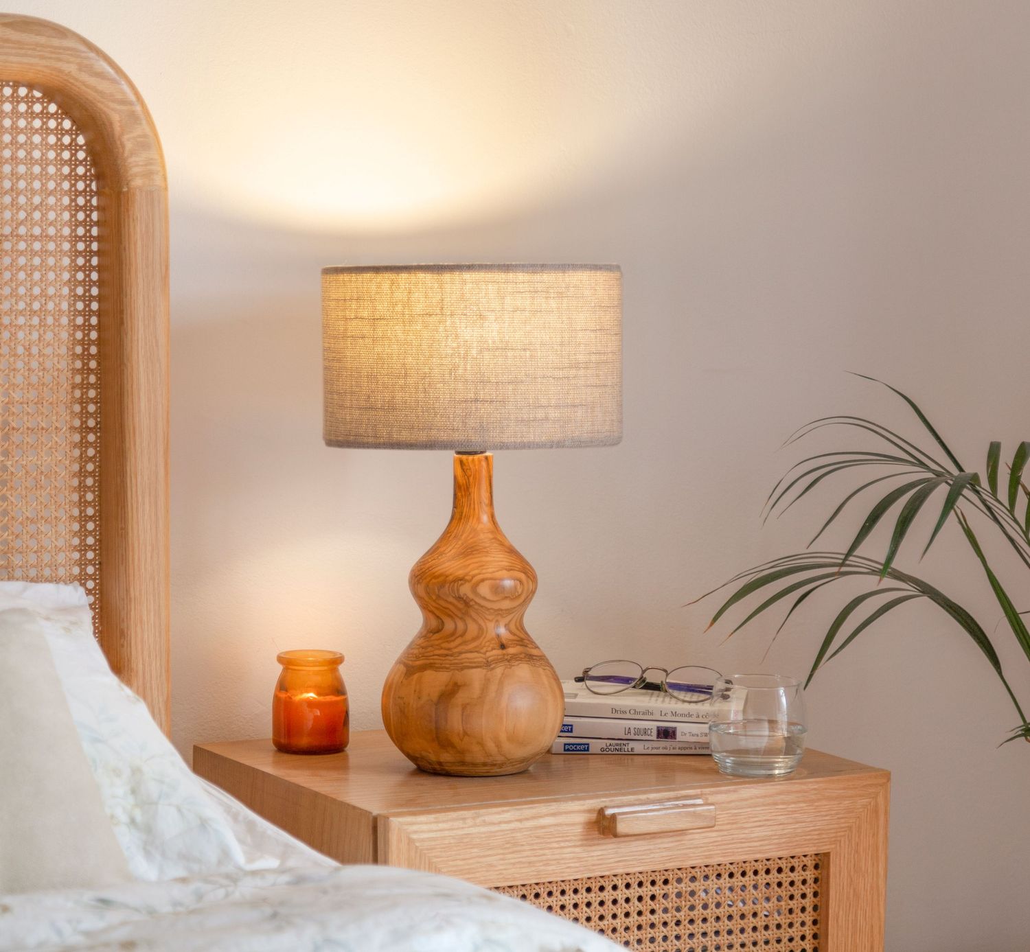 Cozy bedside table with wooden gourd lamp, candle, and palm plant against a neutral wall.