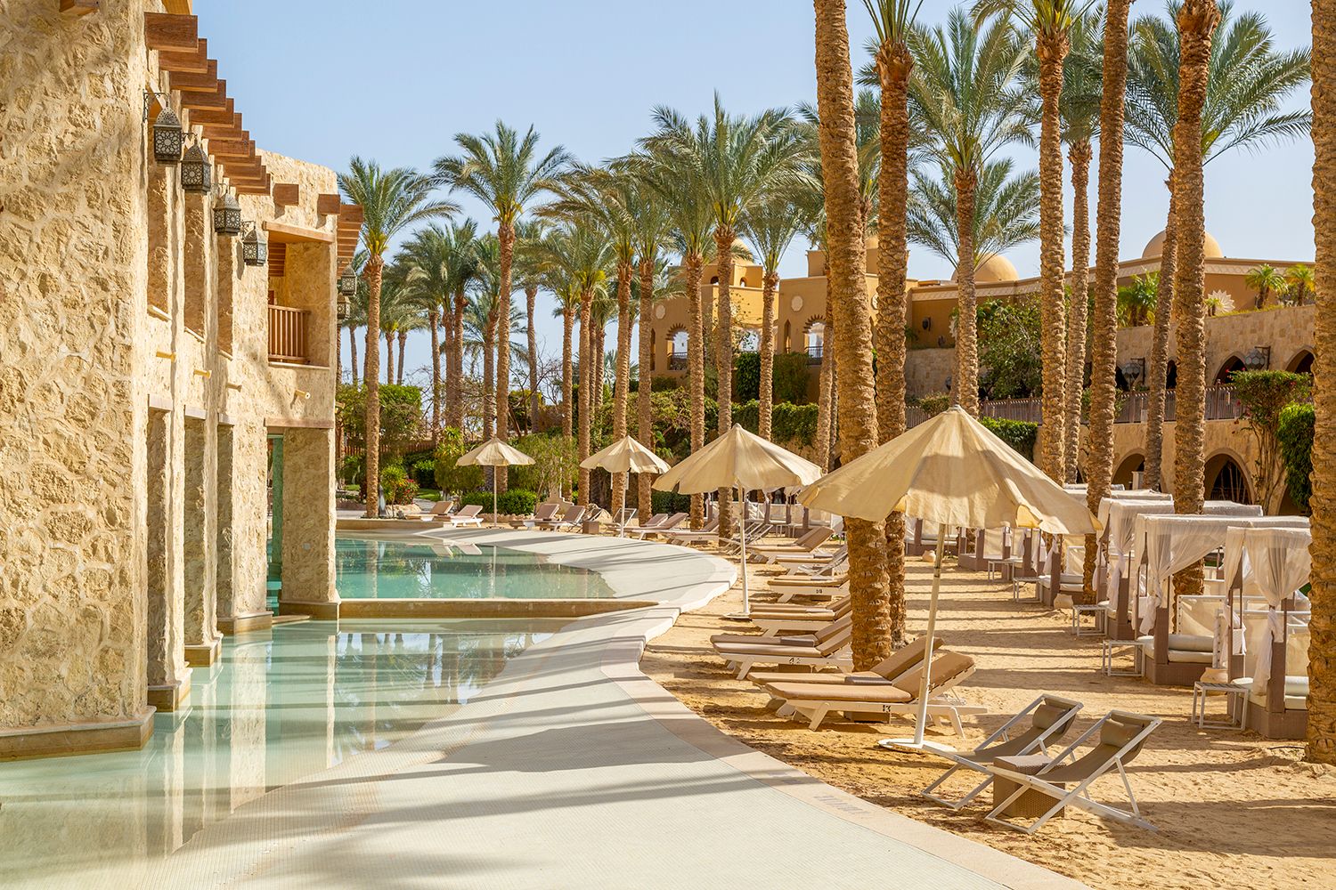 Resort swimming pool lined with palm trees and lounge chairs under thatched umbrellas.