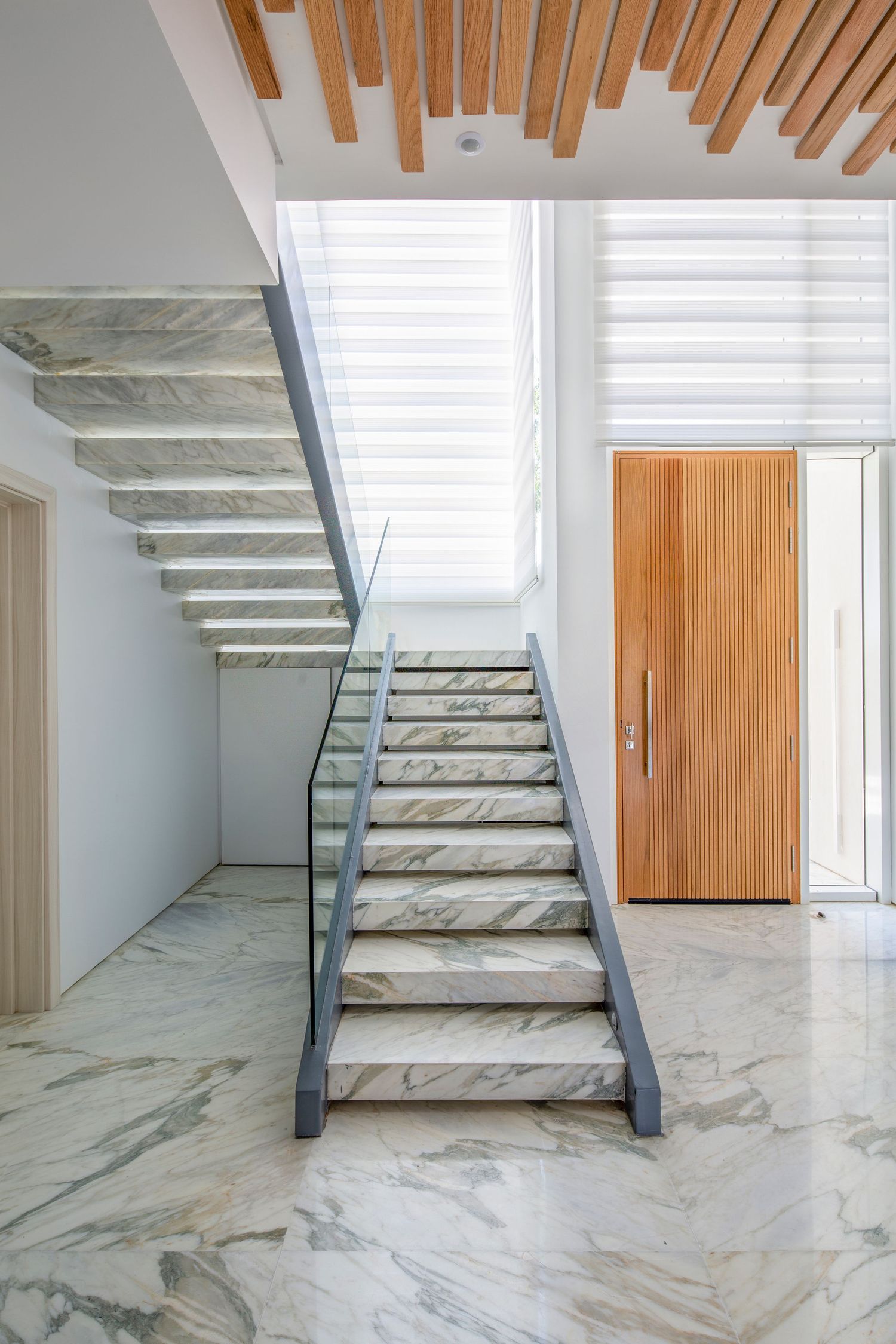 Modern marble staircase with glass railing and wooden ceiling slats in a bright white interior.