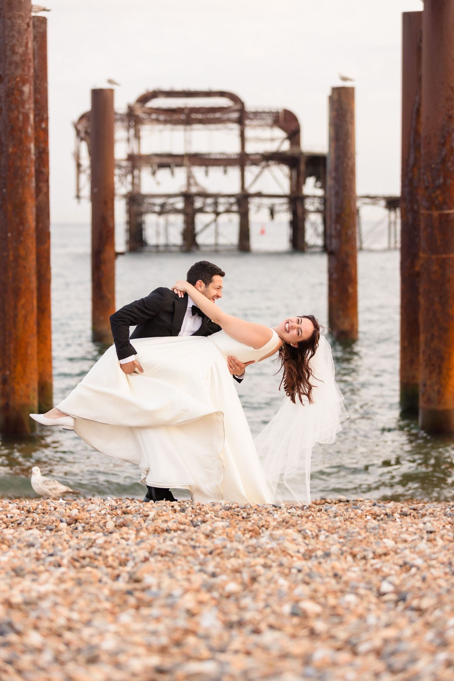 A romantic beach wedding photo with an old pier structure in the background at sunset.