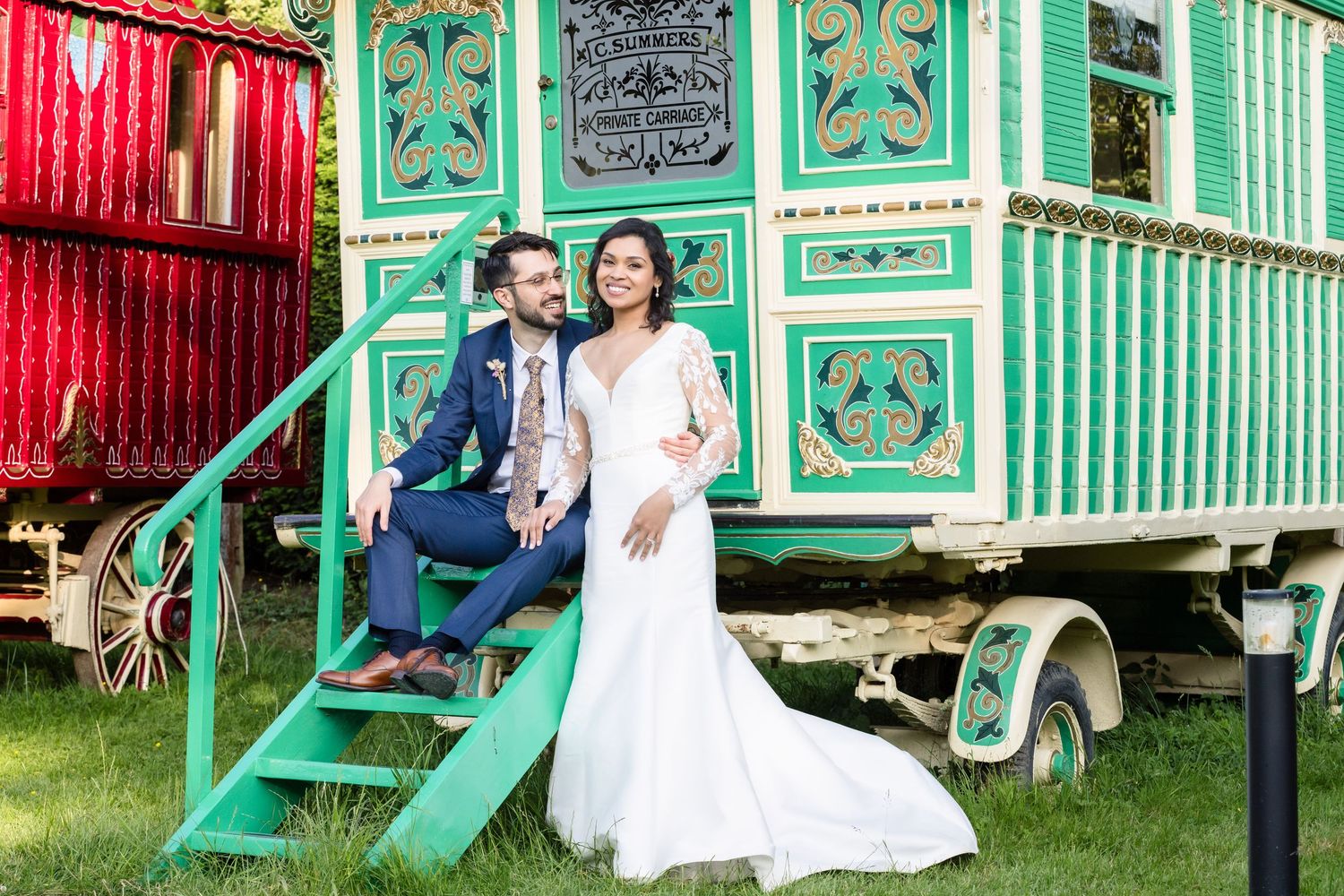 Couple poses together near colorful vintage gypsy caravans decorated in turquoise and red paint at an outdoor event.