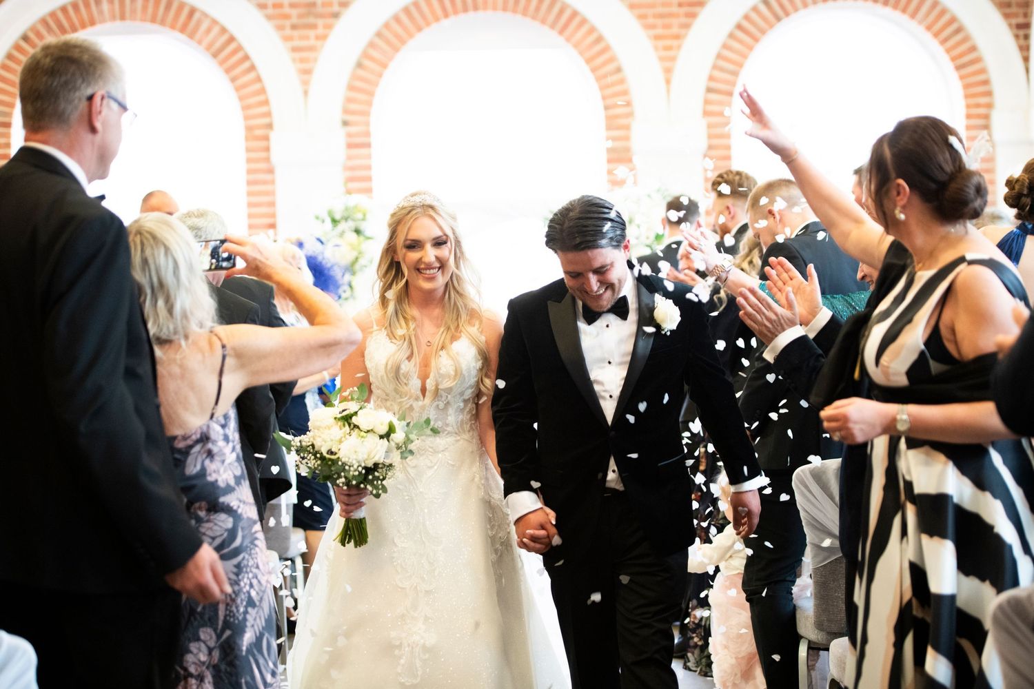 A newlywed couple walks through a shower of white confetti as guests celebrate under brick archways at a wedding ceremony.