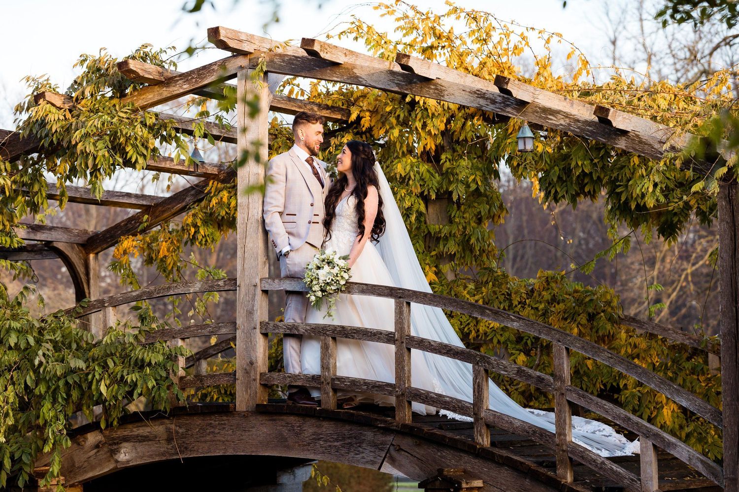 Person in white dress standing on rustic wooden bridge surrounded by autumn trees in garden setting.