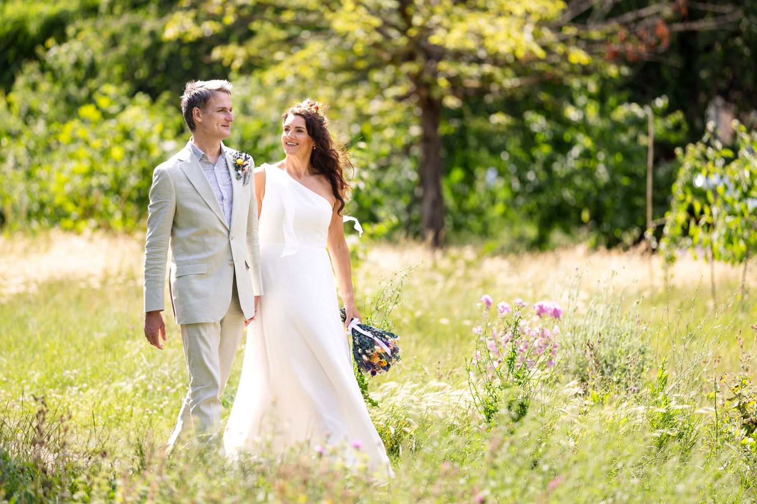 A couple in wedding attire walk together through a sun-drenched meadow with wildflowers and trees in the background.