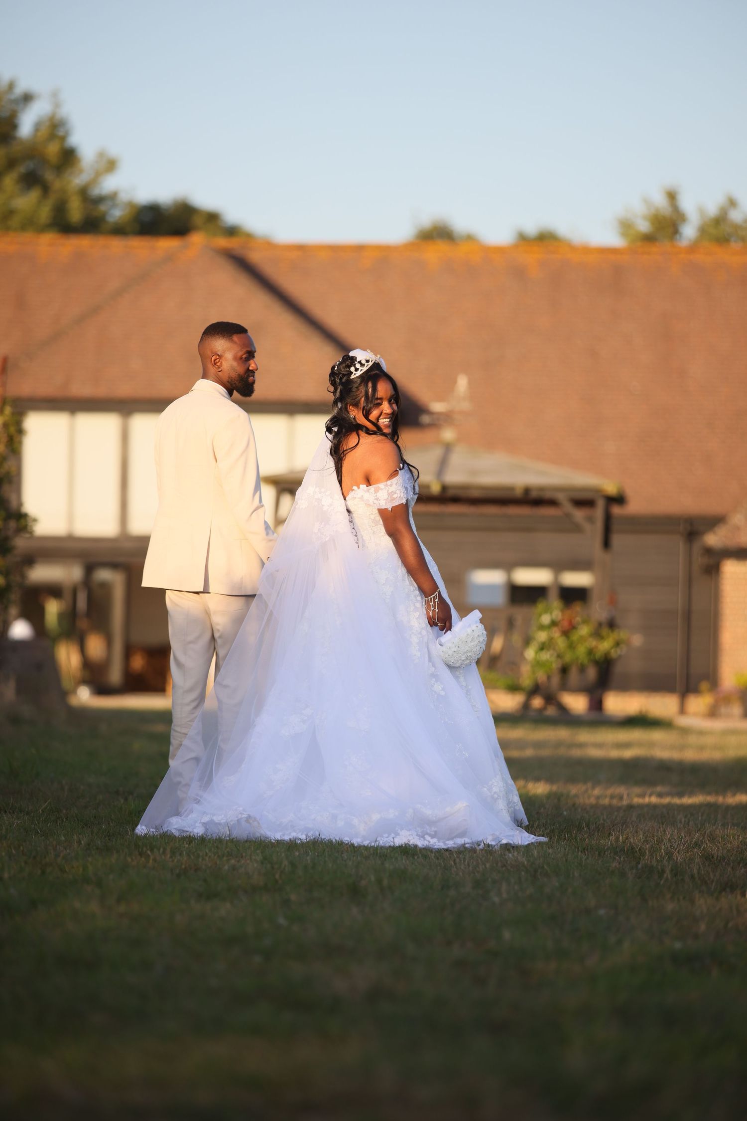 A bride in a puffy white wedding dress walks alongside someone across a grassy field at sunset.