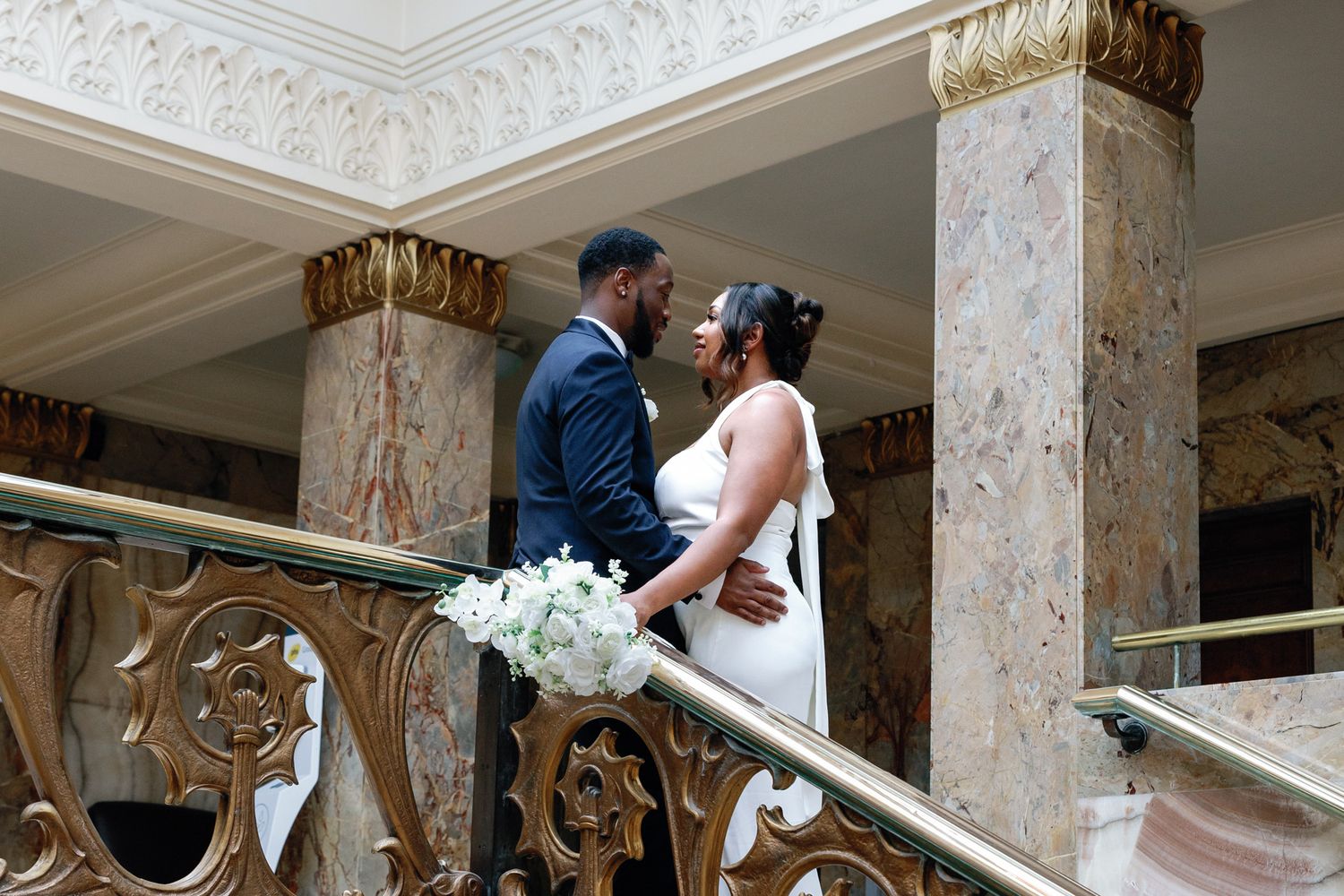 A bride and groom share an intimate moment on an ornate marble staircase with decorative gold railings.