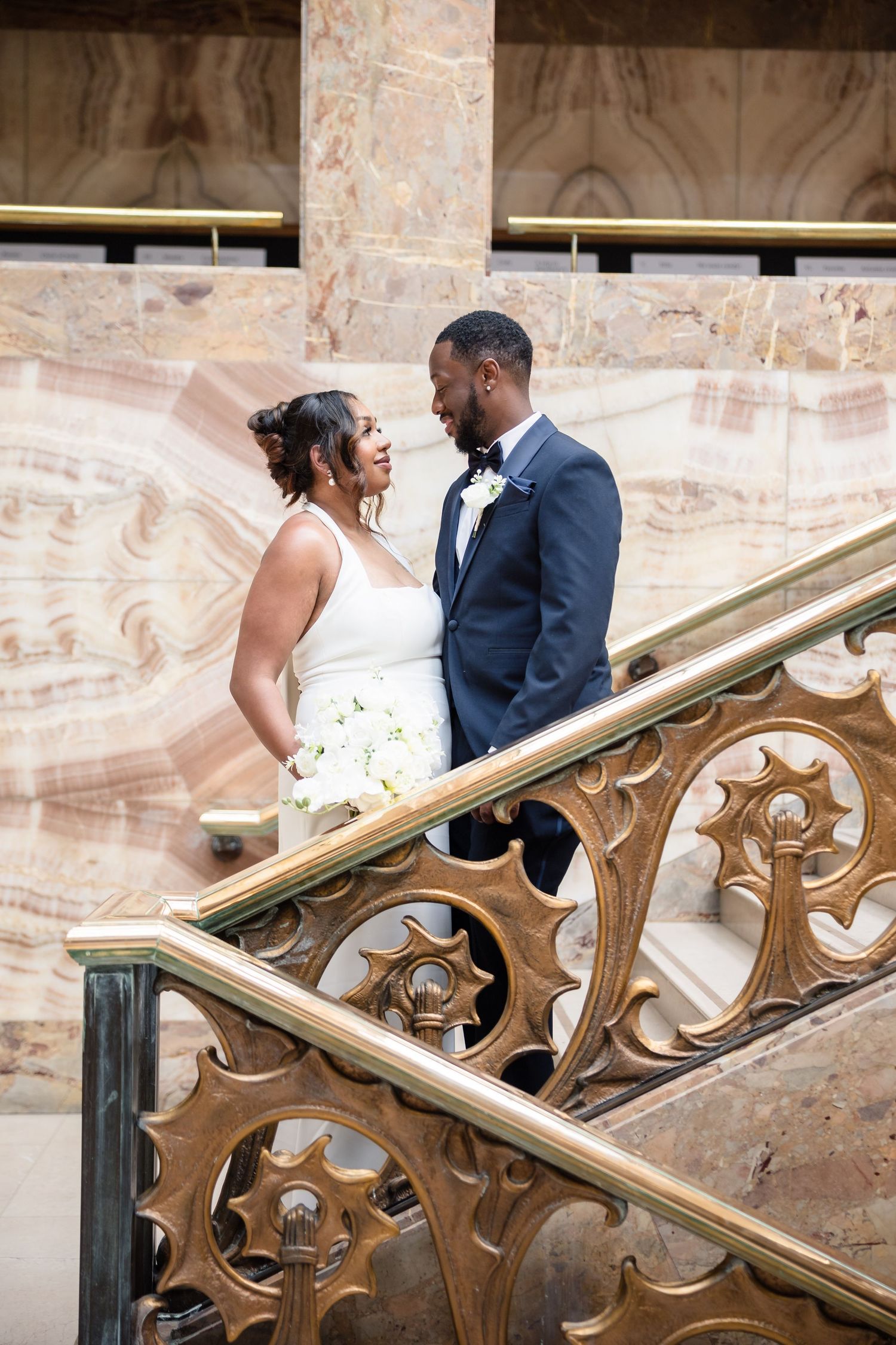 Romantic wedding couple shares intimate moment by ornate brass railing in historic stone building.