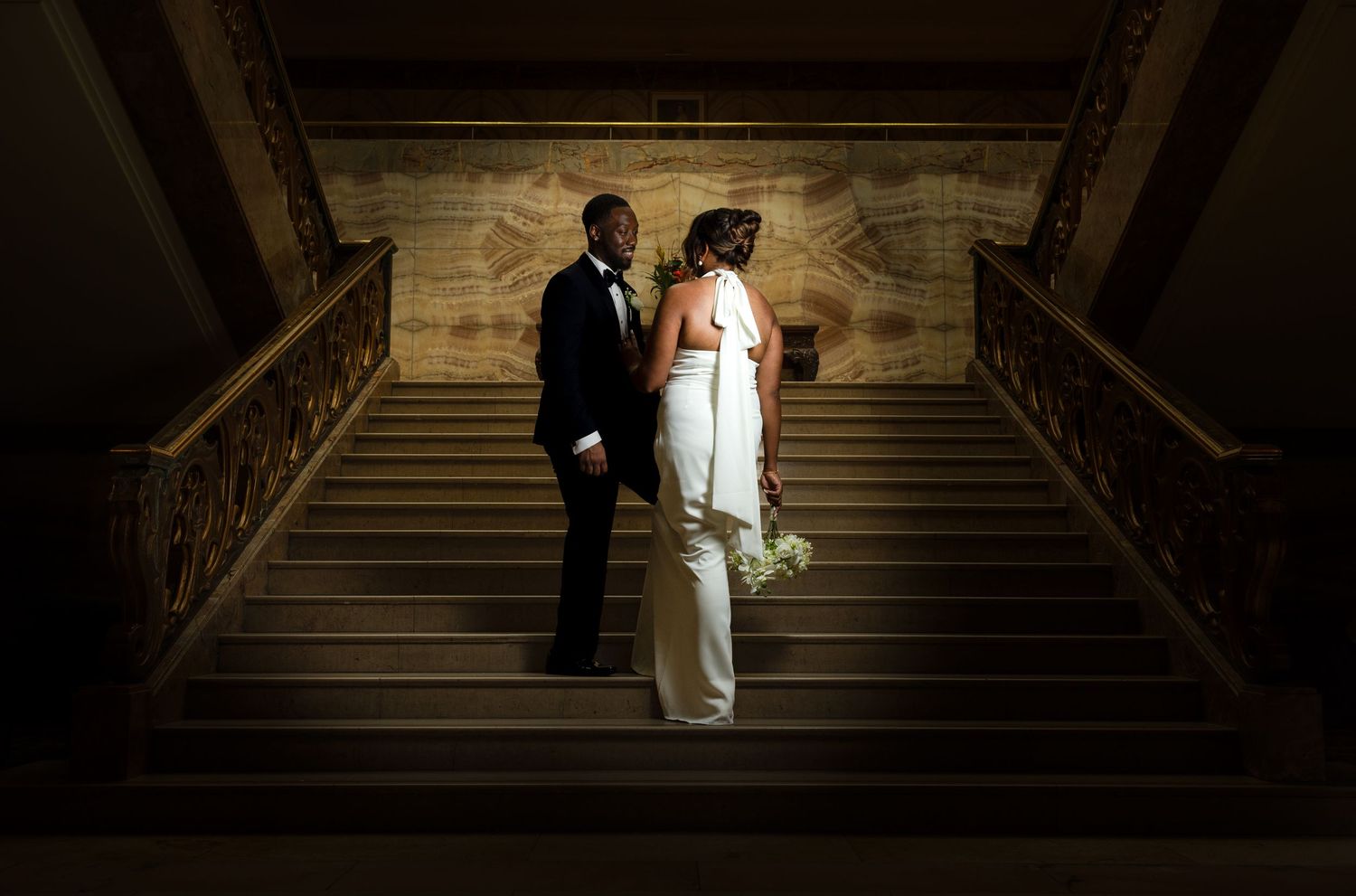 A bride and groom stand on an elegant wooden staircase dramatically lit from above in a dark interior setting.