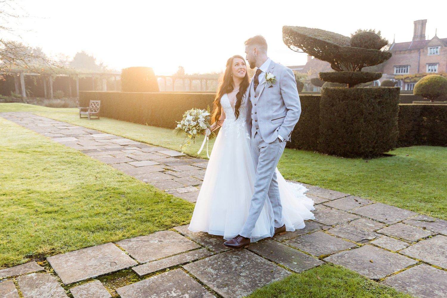 Two people in elegant wedding attire walk along a stone path as golden sunlight streams through trees at sunset.