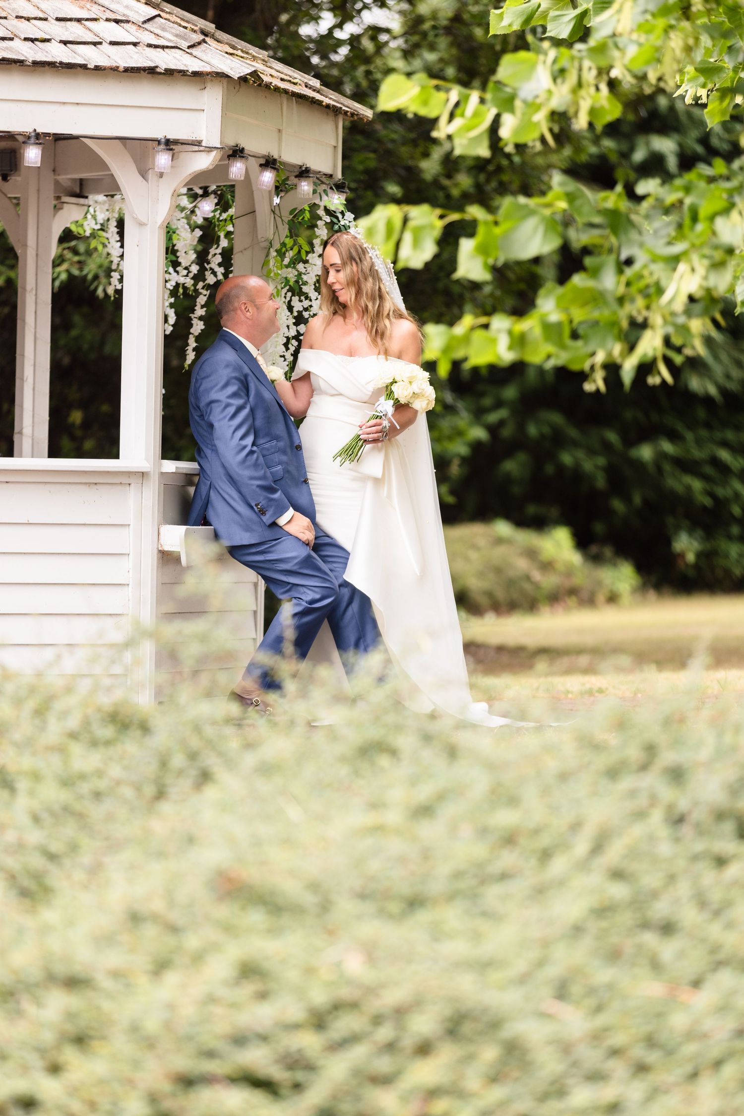 A couple shares a romantic moment in a white gazebo surrounded by lush green foliage during their summer wedding photoshoot.