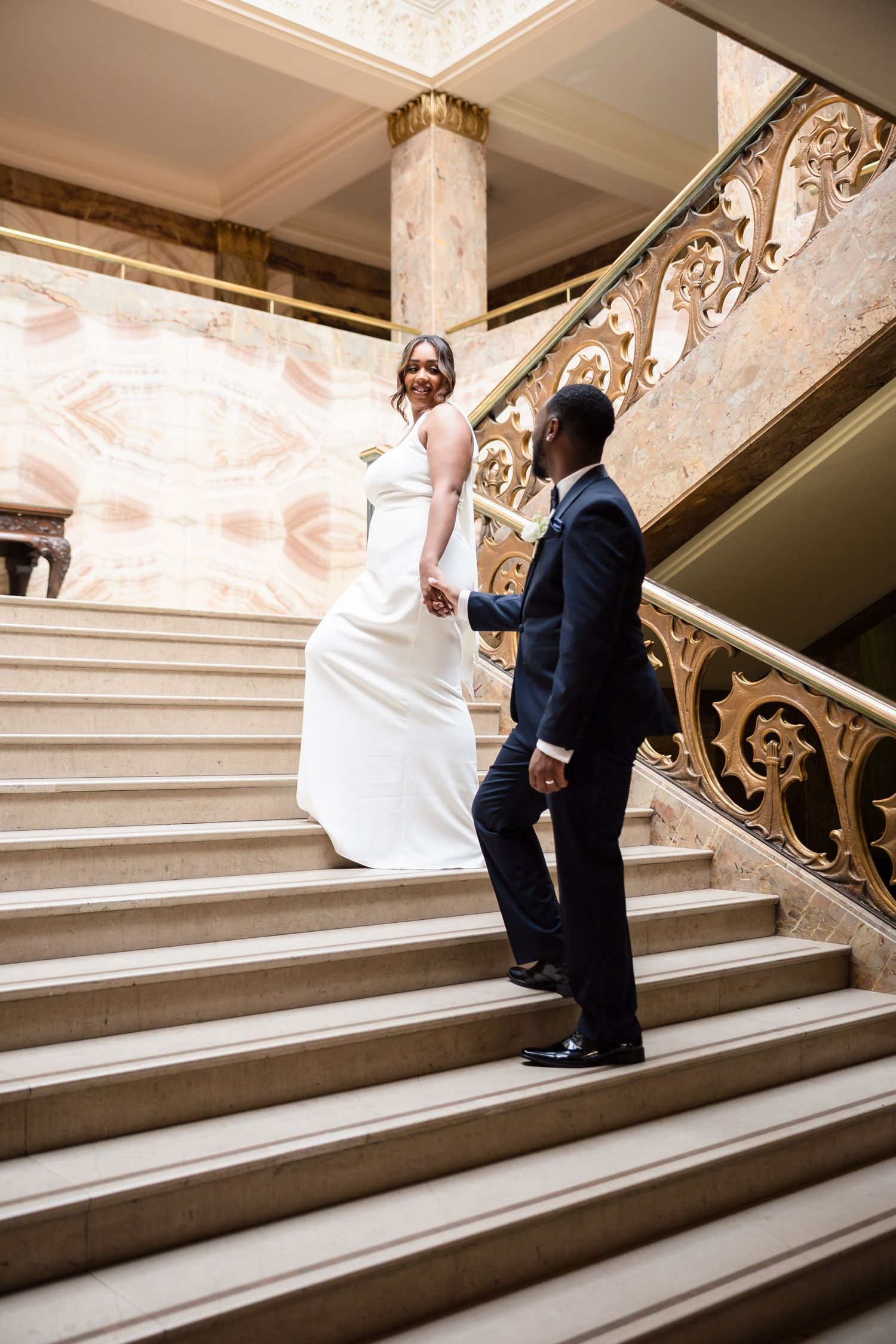 A couple in formal wedding attire walking up an elegant marble staircase with ornate gold railings.