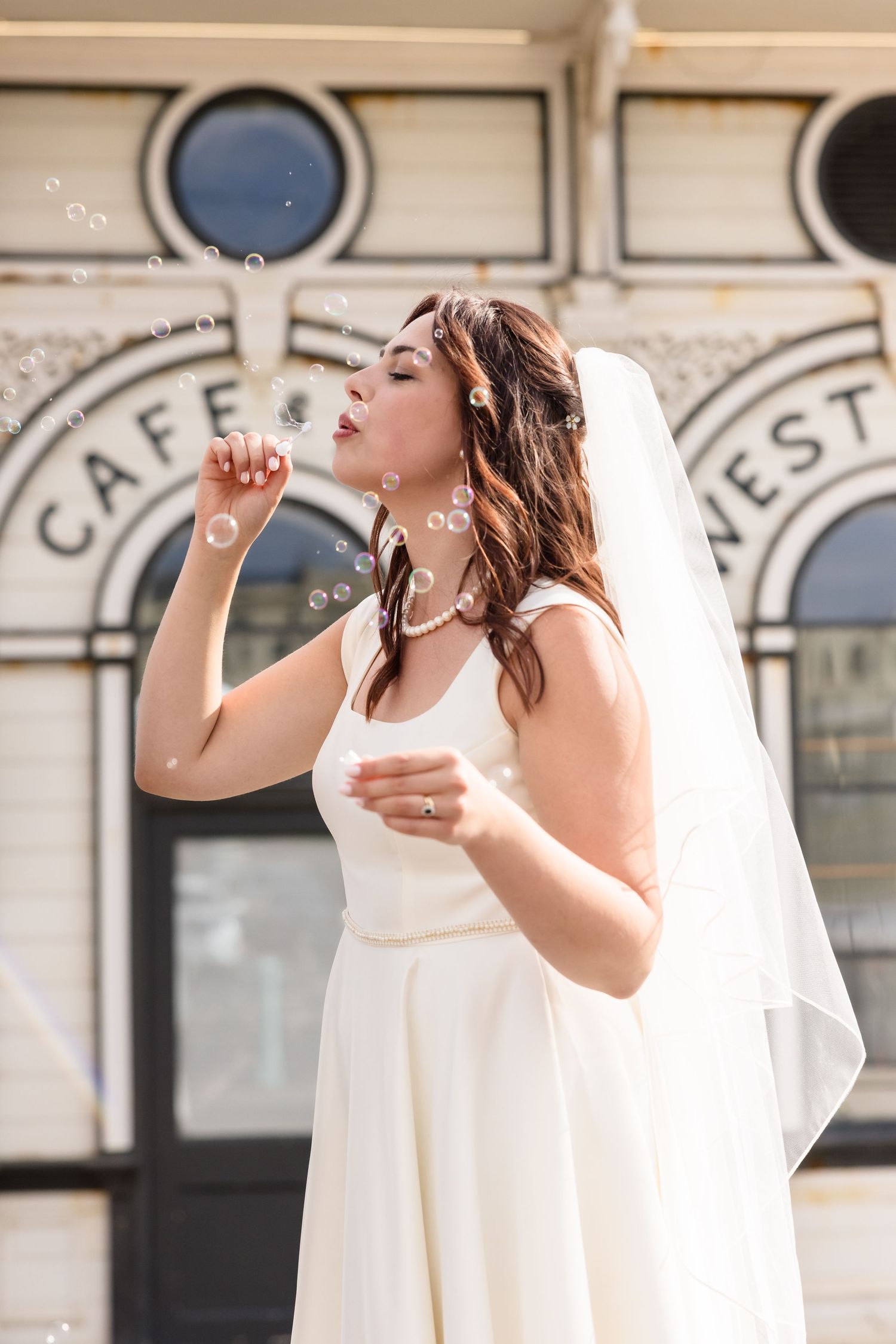 A bride in a white wedding dress blowing bubbles outside a cafe with decorative arches in the background.