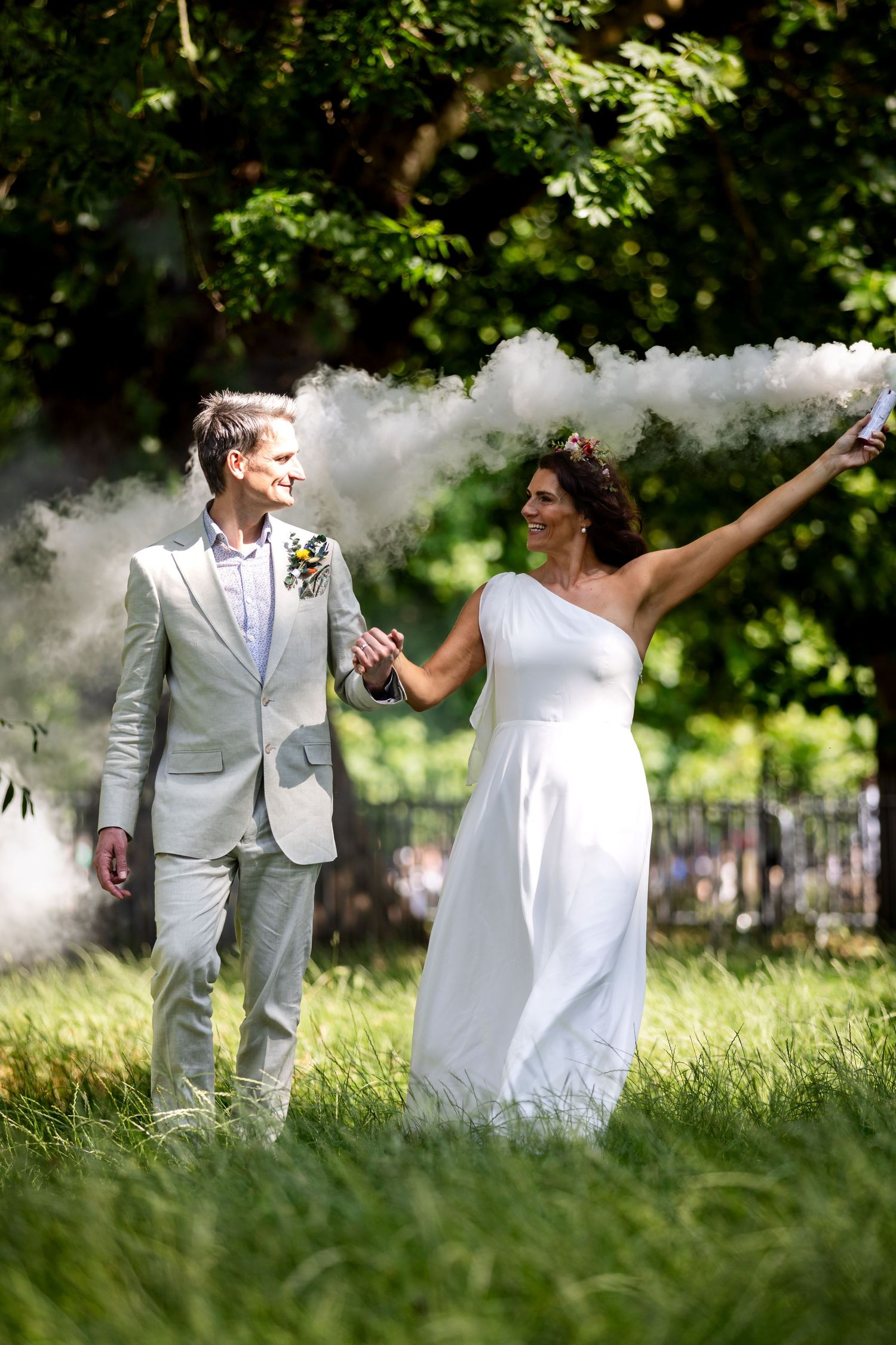 A couple in white wedding attire playfully sprays something in a grassy outdoor setting with trees in the background.