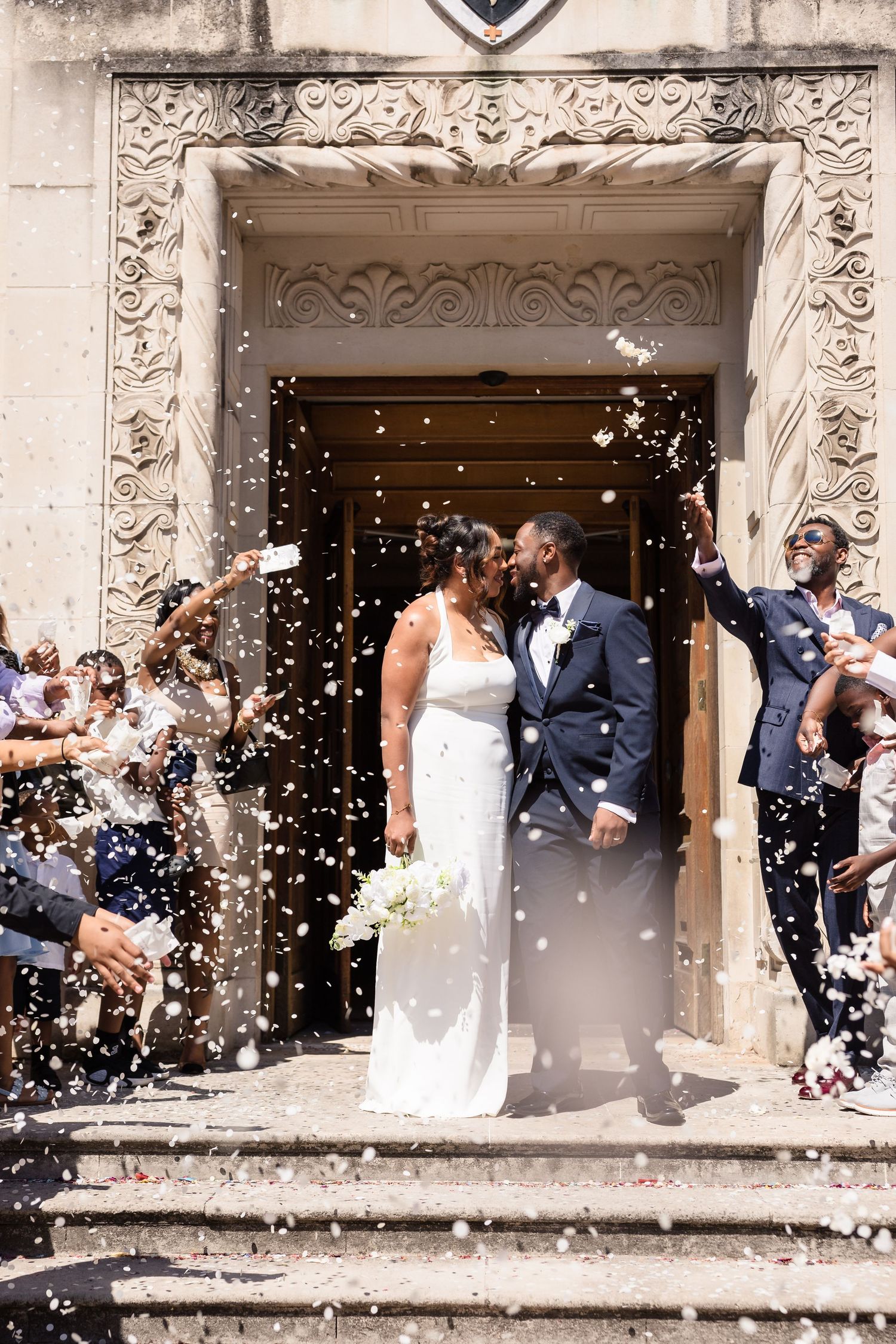 Wedding couple sharing romantic kisses as guests shower them with confetti outside an ornate stone building entrance.