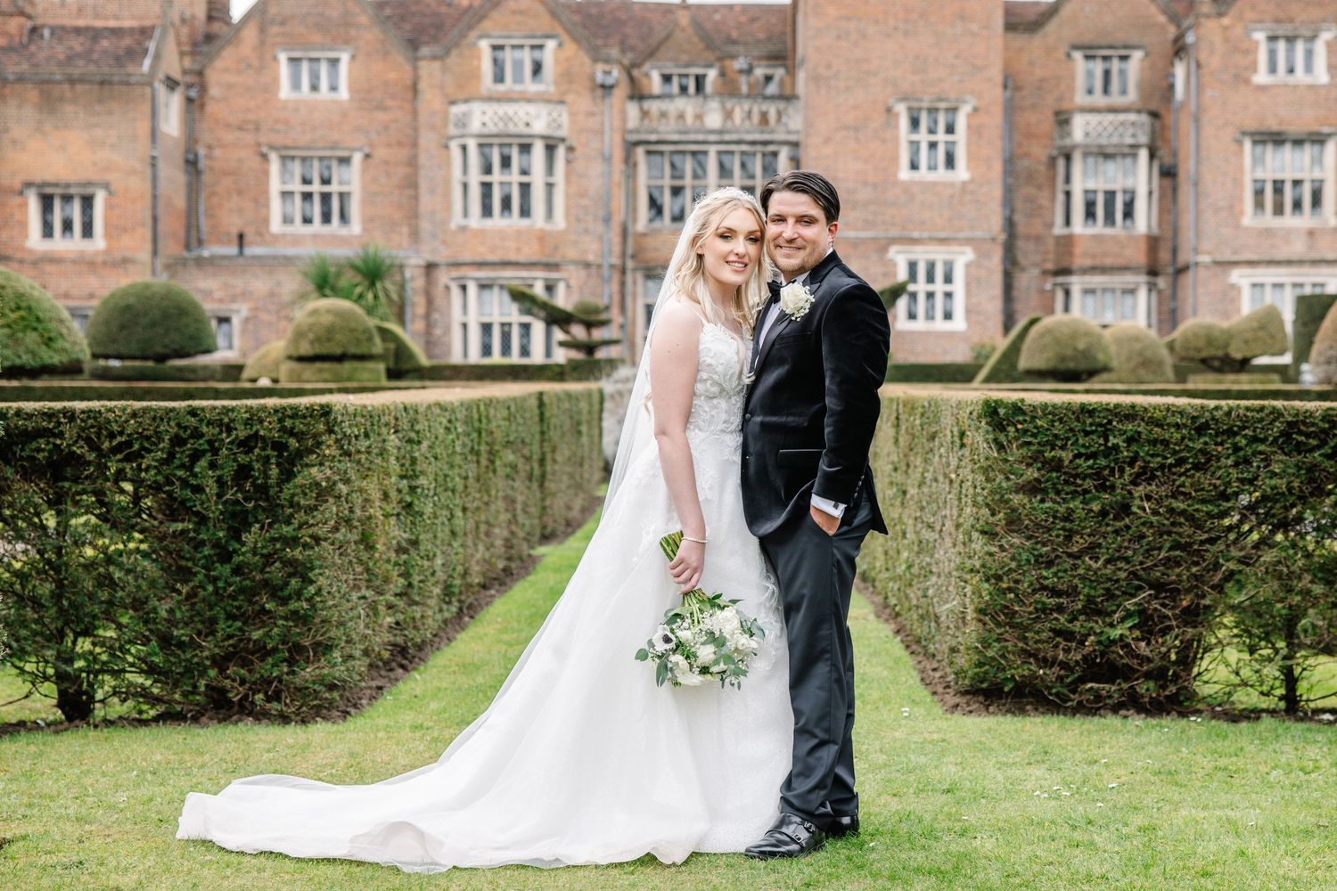 Couple in wedding attire embraces lovingly in a manicured garden with brick buildings in the background.