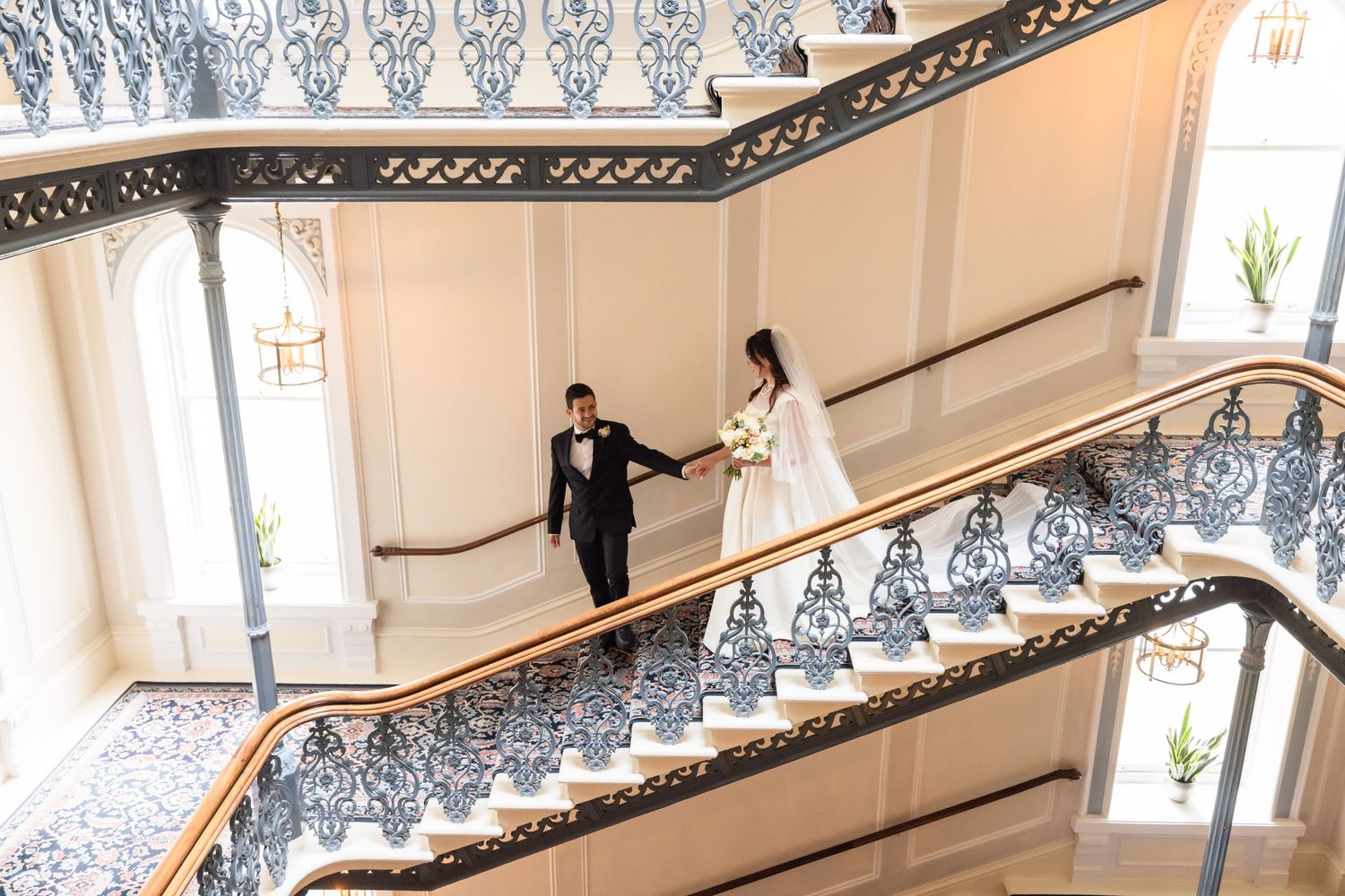 Couple walks down an elegant staircase with ornate black and white railings in a luxury hotel building.