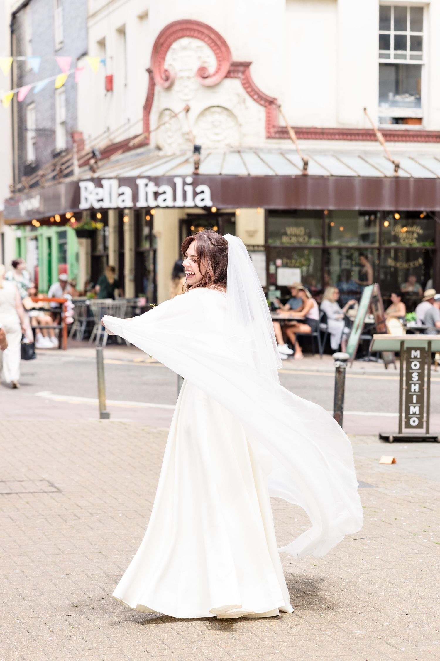 A person in a flowing white wedding dress twirls on a city street in front of Bella Italia restaurant.