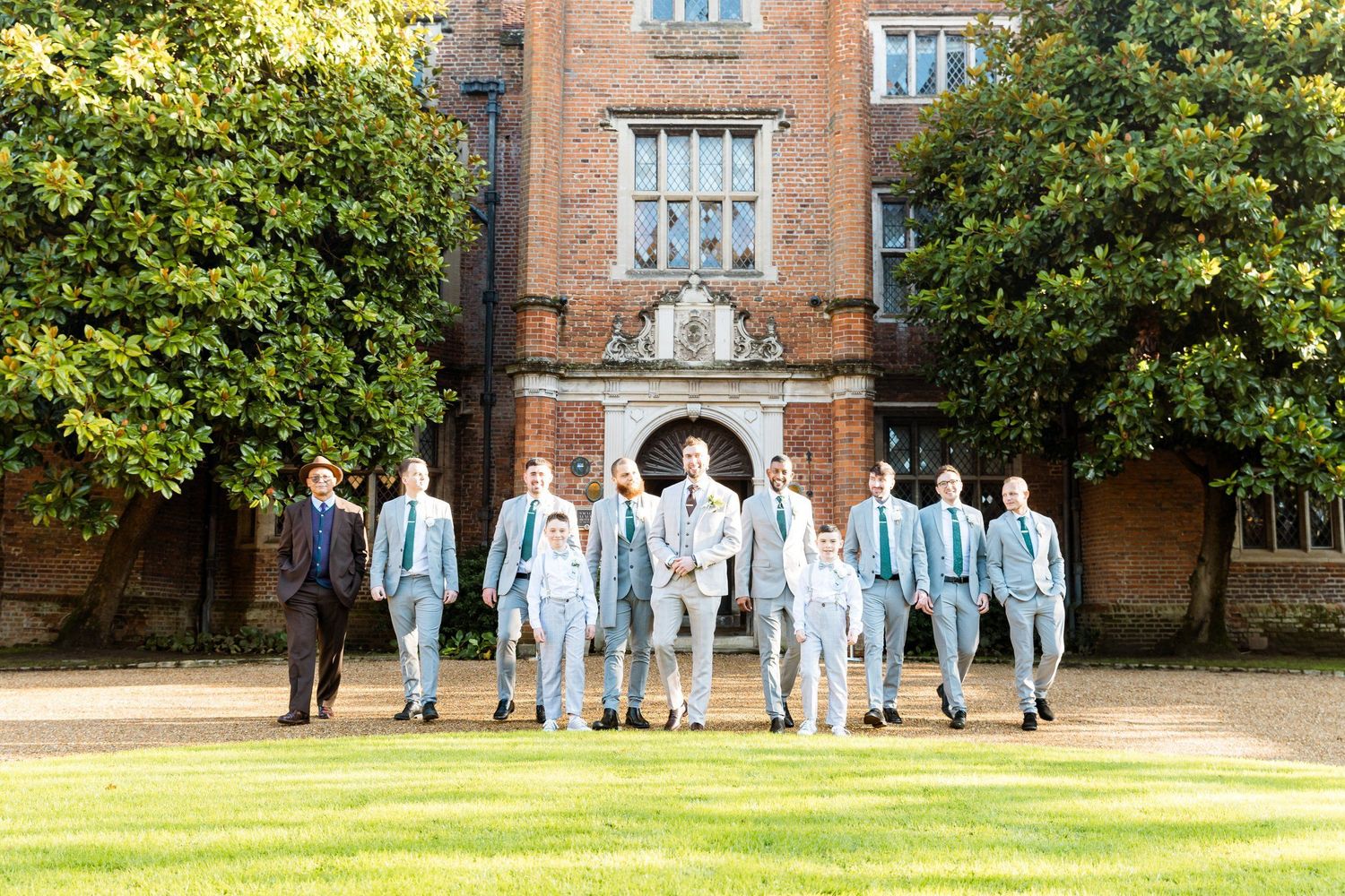 Wedding party wearing matching light blue suits stands in front of a grand brick mansion on a sunny day.